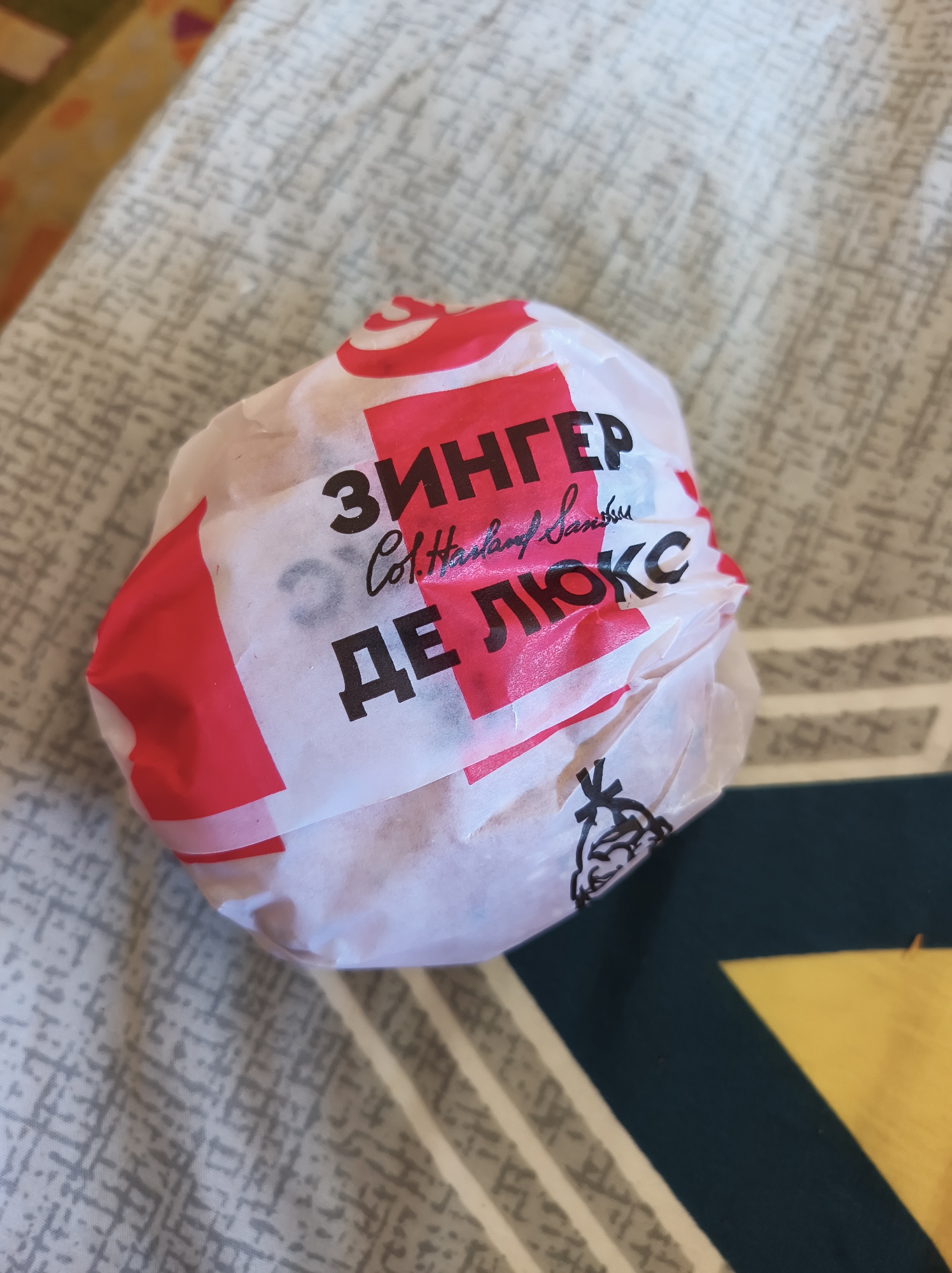 The image shows a wrapped KFC Zinger Burger Deluxe. The wrapper is white with red accents and features the text "ЗИНГЕР ДЕ ЛЮКС" in large black letters, along with some smaller text in script font that is partially obscured. The burger is resting on a textured fabric surface, likely a bedspread or blanket. The fabric has a pattern of gray and white lines and shapes. To the right of the burger, a portion of another patterned fabric in dark blue and yellow is visible. There is no indication of the location being Baranavichy, Belarus. The image is a close-up shot, focusing on the food item.
