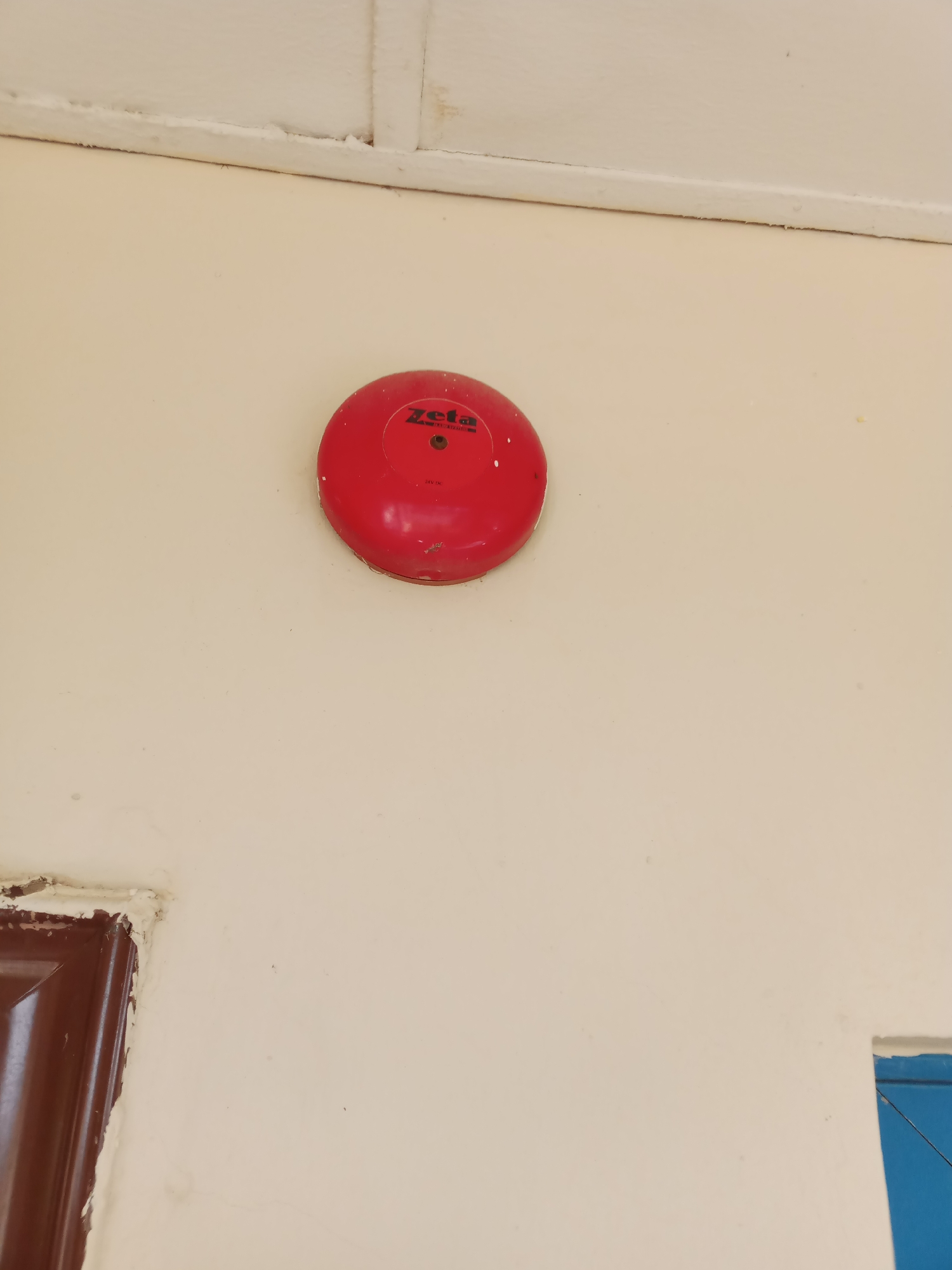 A red Zeta brand alarm bell is mounted on a cream-colored wall. The alarm bell is circular with a raised center, and the "Zeta" logo is visible in black text. Below the logo, "24V DC" is also printed. The wall appears to be part of an interior space, possibly an office or a common area in Jalingo, Nigeria. The ceiling above the wall is white with a visible seam. In the bottom left corner of the image, a portion of a dark brown door frame is visible, showing some signs of wear around the edges. In the bottom right corner, a sliver of a blue frame or object is present. The lighting suggests it is daytime.