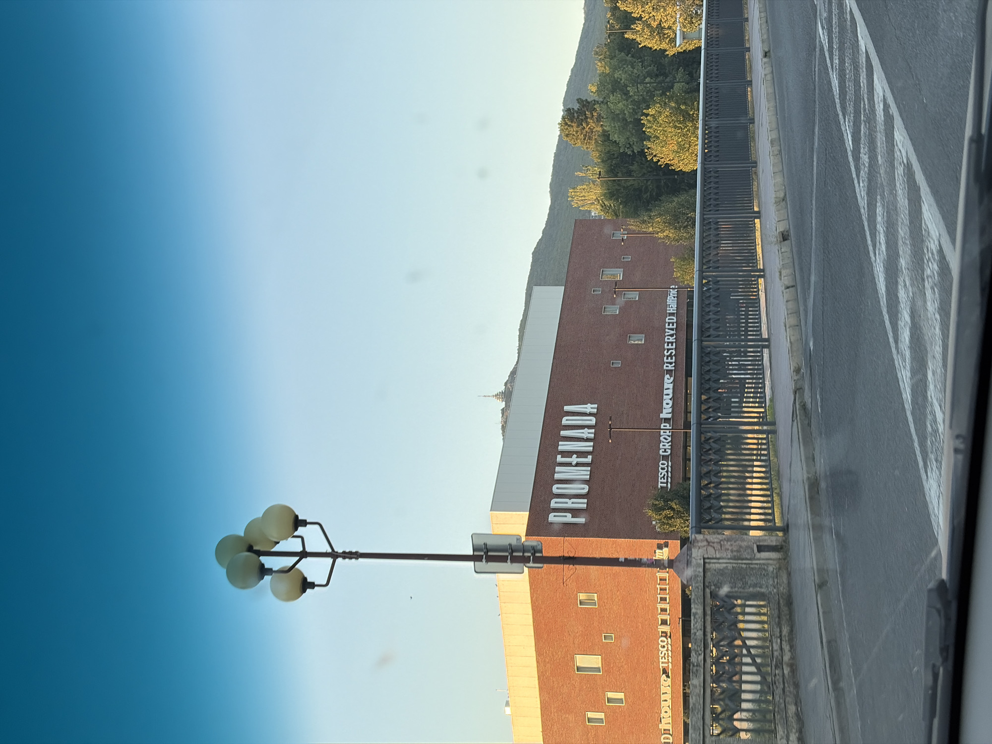 The image captures a view of the Promenada shopping center in Nitra, Slovakia, as seen from a street. The shopping center is a large brick building with white lettering spelling out "PROMENADA" across its facade. Above the brickwork, there is a section of white cladding. Several small, square windows are visible on the brick part of the building. Below the "PROMENADA" sign, there are signs for "TESCo" and "RESERVED HalfPrice".
In the foreground, a street is visible with a pedestrian crossing marked by white stripes. A sidewalk and a decorative metal railing of a bridge or walkway are present between the street and the shopping center. A street lamp with multiple globes stands tall to the left of the shopping center.
The background features lush green trees and a hillside with a prominent structure, possibly a church or monument, on its peak. The sky is a clear, bright blue, indicating fair weather, and the light suggests it is either early morning or late afternoon. There are no people visible in the image.