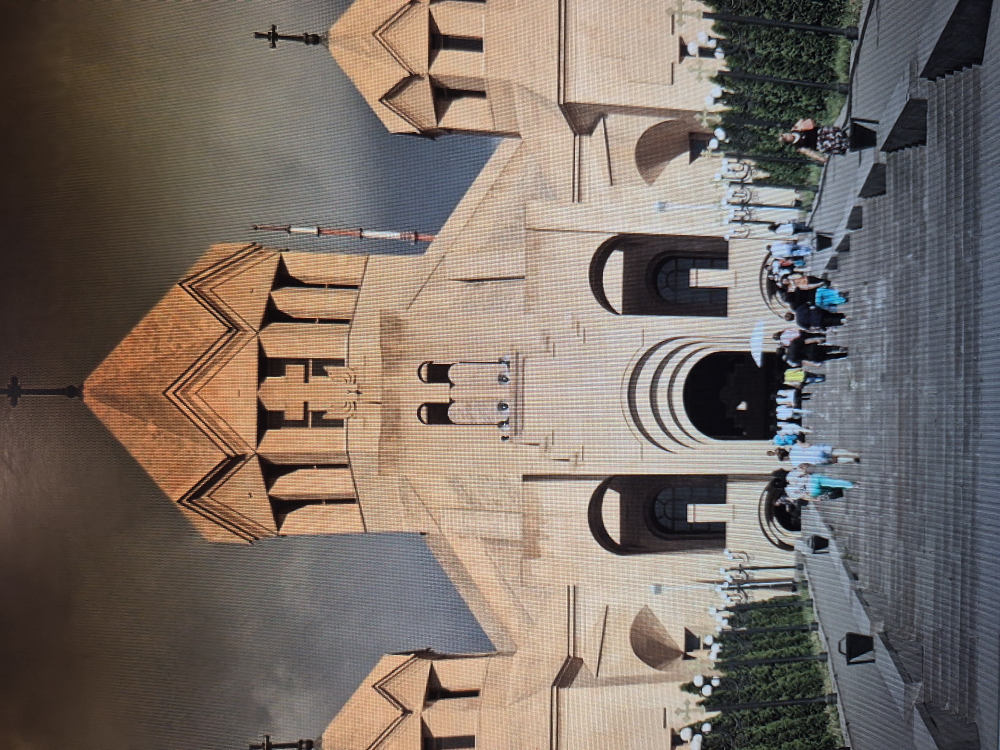 The image shows St. Gregory the Illuminator Cathedral in Yerevan, Armenia. The photograph is presented at an unusual angle, appearing almost like a mirrored reflection. The architecture of the cathedral is prominent, featuring light-colored stone and geometric patterns. A group of people is visible on the steps leading up to the entrance of the cathedral. Manicured landscaping, including rows of shrubs, line the walkway. A grey sky suggests a cloudy day.