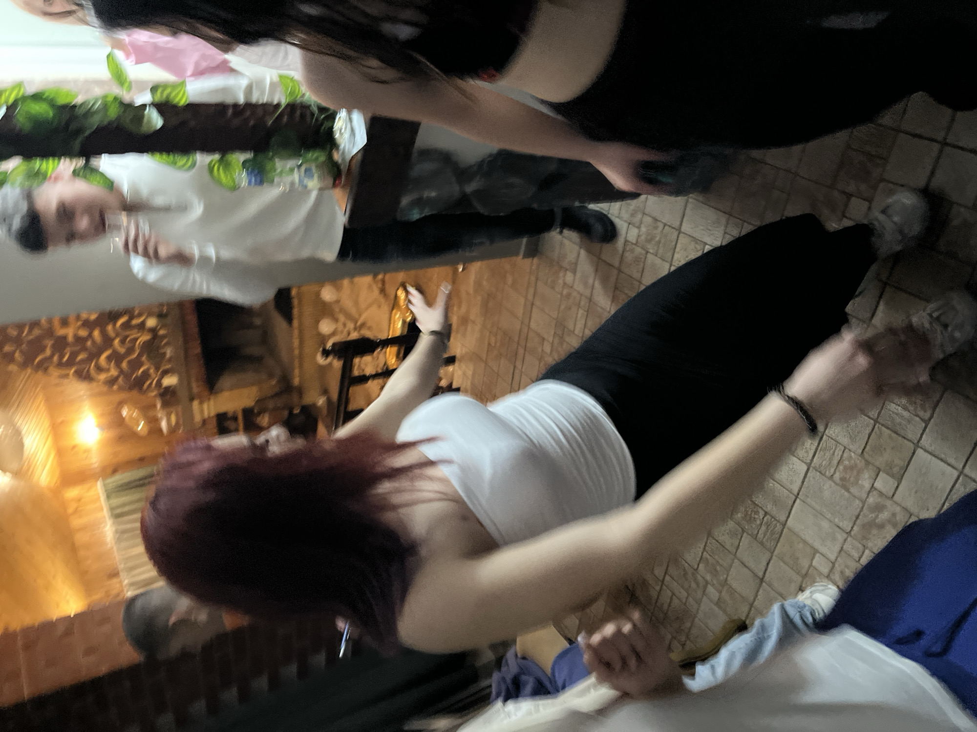The image captures a blurred scene, likely indoors, with multiple individuals present. A woman with dark red hair wearing a white tank top and black leggings is at the forefront, her arms extended. Another woman wearing black top and leggings is present. A man in a white shirt and dark pants stands near a table with plants. The surroundings appear to be a dining area, characterized by tile flooring and wooden wall paneling with decorative light fixtures. The exact location within Presoka, Bulgaria, cannot be determined based on the provided image.