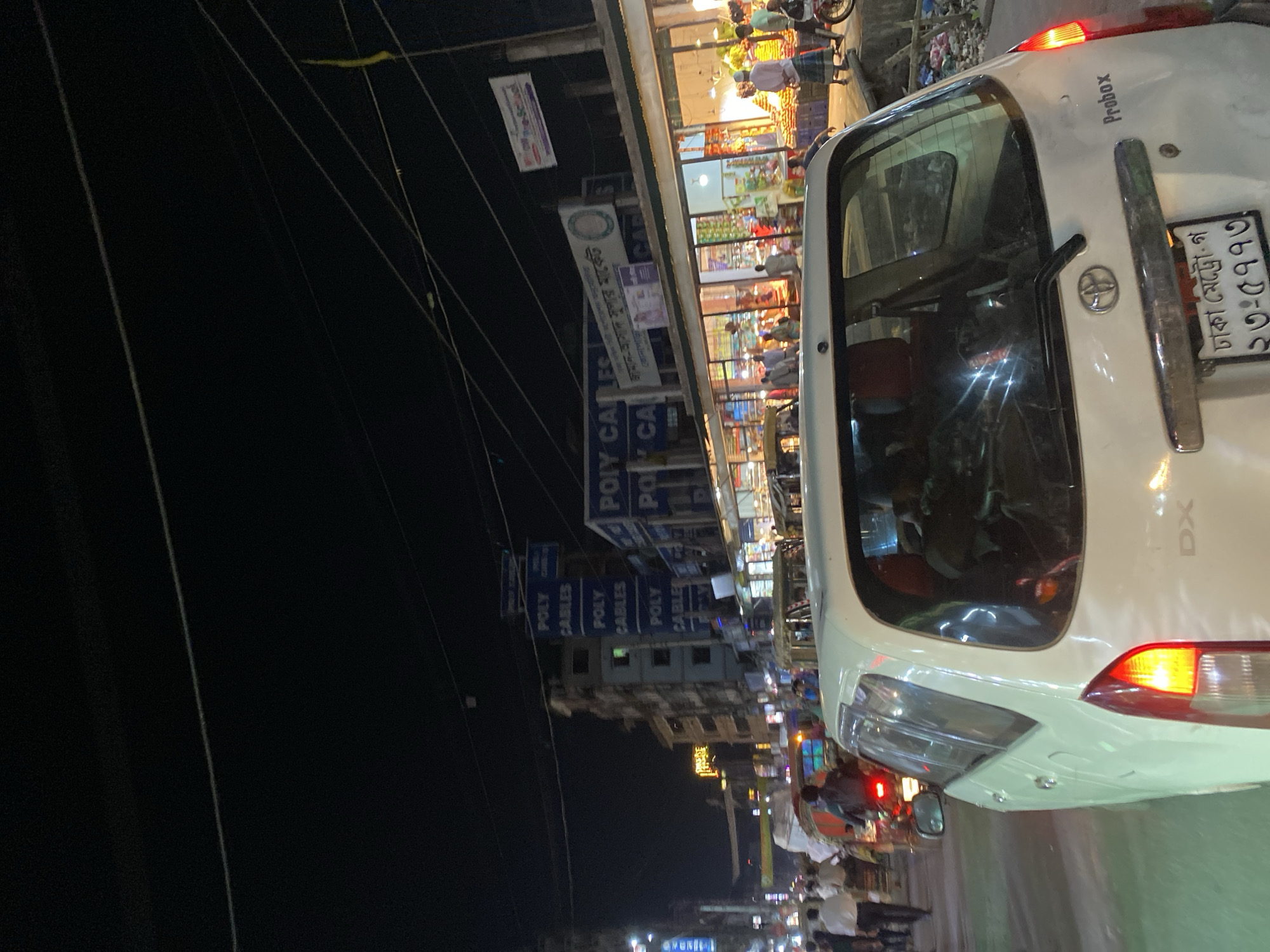 The image depicts a vibrant night street scene in Dhaka, Bangladesh, bustling with activity. In the immediate foreground, the rear passenger side of a white Toyota Probox car is visible, identified by its "Probox" and "DX" badging, and a Toyota logo. Its taillights are illuminated, and the license plate clearly reads "ঢাকা মেট্রো-গ ২৩-৫৭৭৩" (Dhaka Metro-G 23-5773).
Beyond the car, the street is lined with numerous shops and market stalls, brightly lit against the dark night sky. Many people are present, moving along the street, shopping, or tending to their businesses, although their figures are somewhat blurred due to motion or low light conditions. The scene suggests a busy commercial area.
Overhead, a complex network of power lines is strung across the frame. Several shop signs are discernible: prominent blue signs display "POLY CABLES" in English, suggesting an electrical supplies store. Another large sign reads "জামান-আসাদ সুপার মার্কেট" (Zaman-Asad Super Market), with "কোনাপাড়া" (Konapara) also visible on a nearby sign, likely indicating a specific market or area. The overall lighting is artificial, coming from the shops and vehicle lights, creating a warm and energetic atmosphere typical of a bustling market at night.