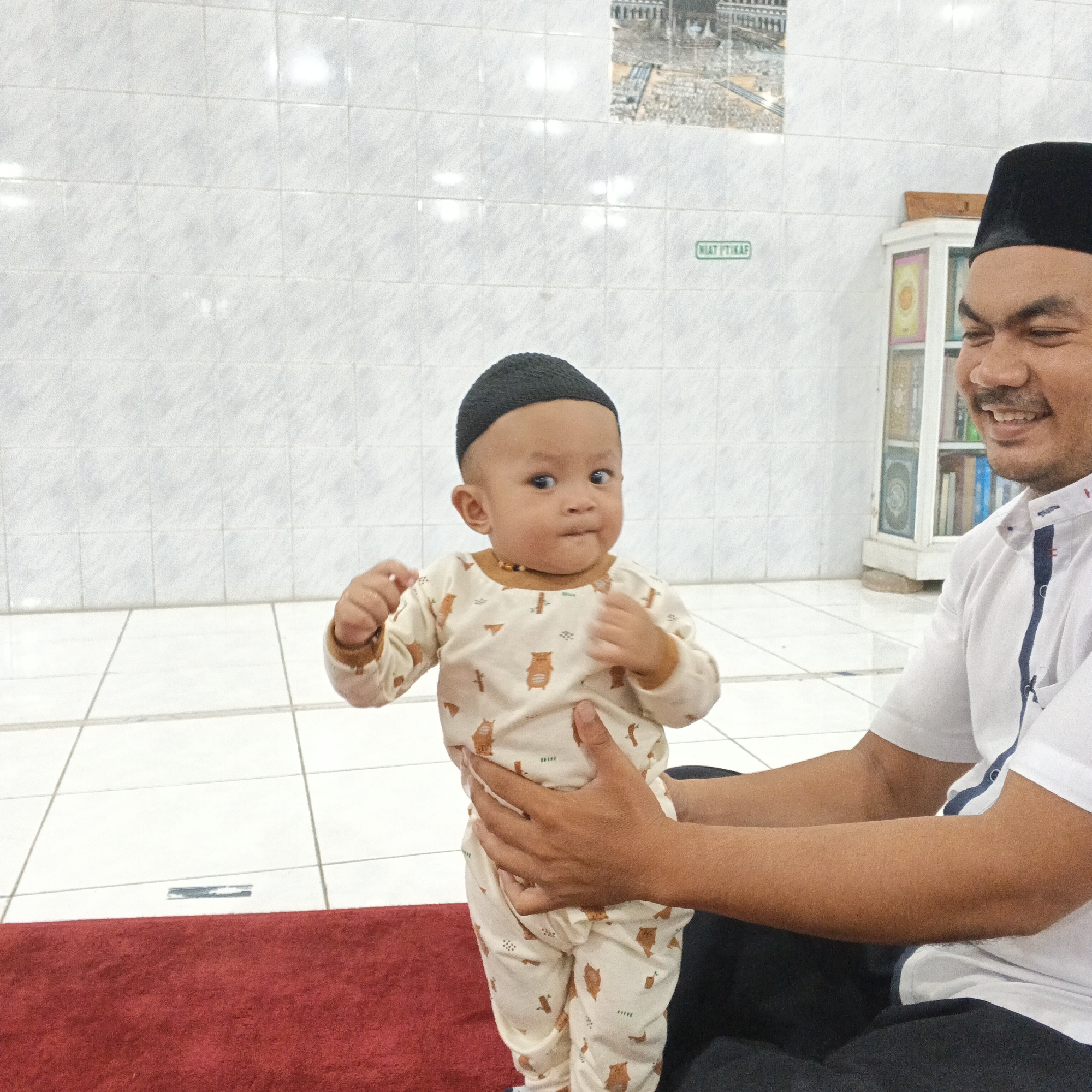 In the city of Meunasah Raya, Indonesia, a man wearing a black peci and a white shirt with a blue stripe down the front is holding a baby. The baby is wearing a beige onesie with a pattern of cartoon bears and is also wearing a black peci. The man is smiling and looking at the baby. The background appears to be an indoor space with white tiled walls and a red carpet. A cabinet with books and other items is visible on the right side of the image. A framed picture of a mosque is on the wall in the background. The overall scene suggests a religious or familial gathering.