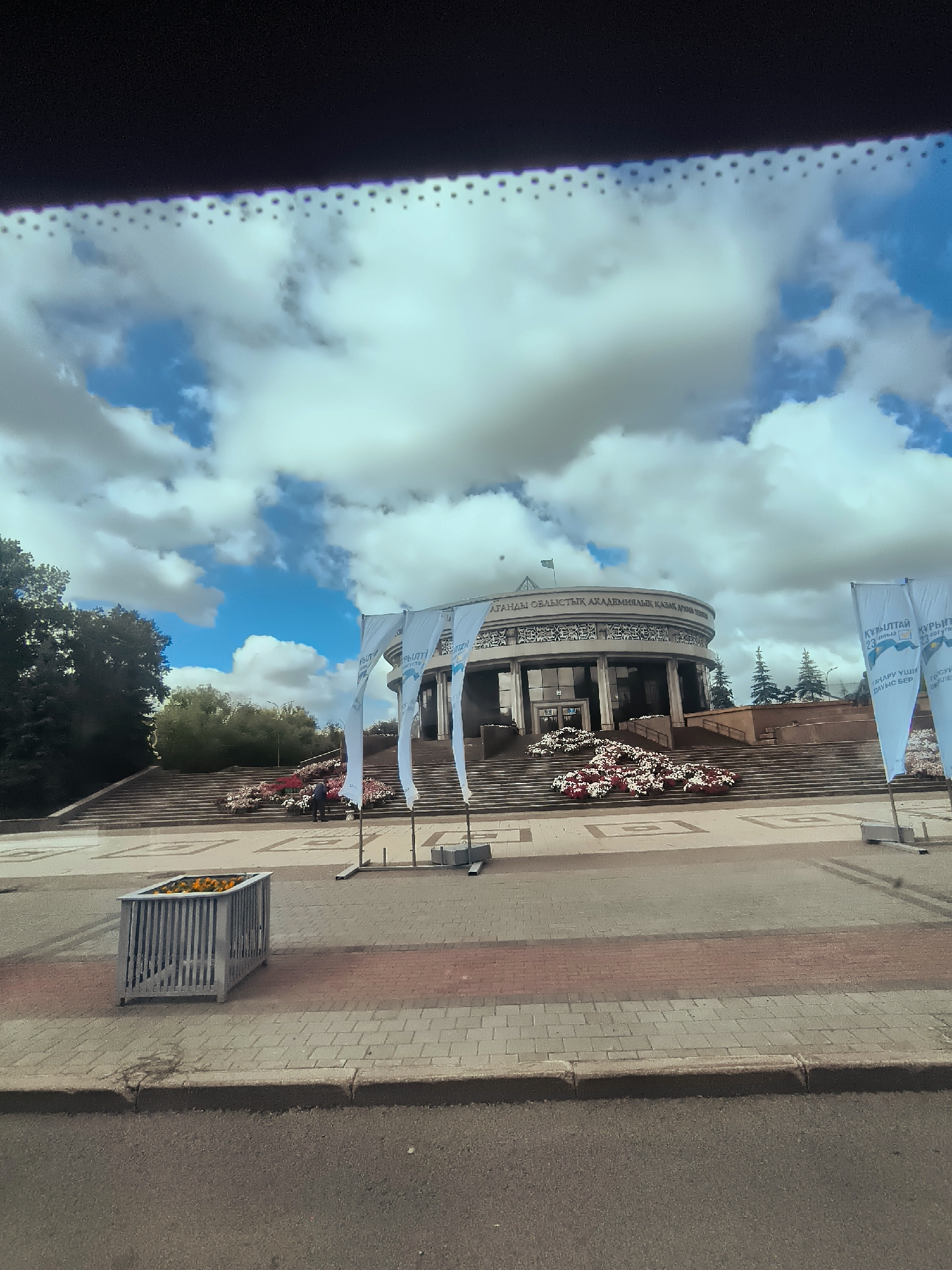 The image shows the city administration building of Qarağandı, Kazakhstan. The building is a circular, modern structure with a wide staircase leading up to the entrance. The staircase is decorated with large floral arrangements. Several white flags with text are displayed in front of the building. The sky is filled with dramatic clouds. In the foreground, there is a metal planter box filled with yellow flowers and a paved walkway. A person is visible on the stairs, appearing to tend to the flowers. The environment is urban, with trees and other buildings visible in the background.