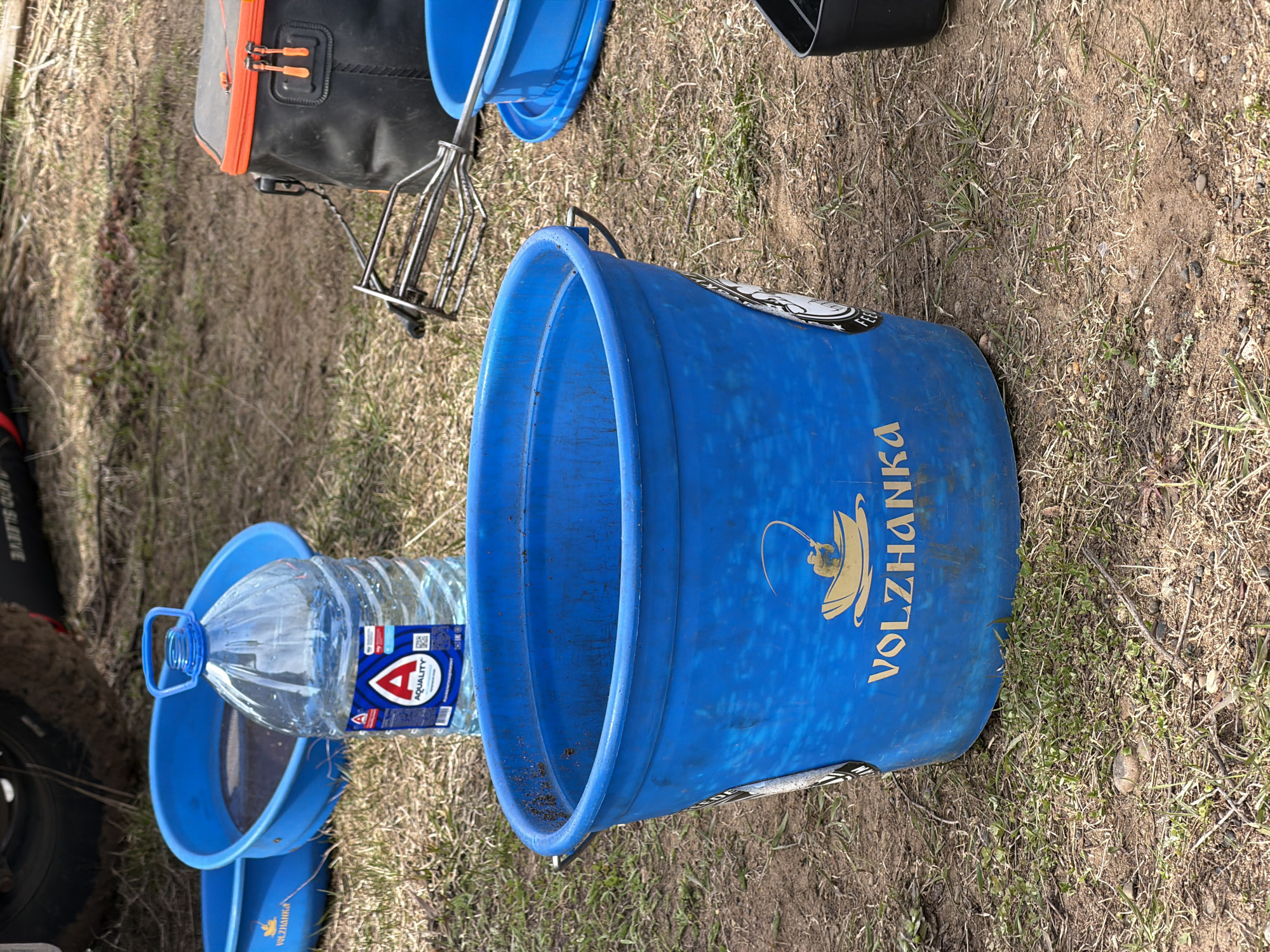 This image captures an outdoor scene featuring an array of fishing equipment laid out on dry, grassy ground in Nikolo-Beryozovka, Russia. The primary subject is a large, vibrant blue plastic bucket, positioned upright, with the brand name "VOLZHANKA" printed vertically in gold lettering on its side. Below the text is a gold-colored logo depicting a person fishing from a small boat. Partially visible on the bucket's side is a sticker with the word "FEED".
Adjacent to the large bucket is a smaller blue basin or sieve, containing a clear plastic water bottle with a blue handle and cap. The bottle's label is clearly visible, displaying the brand "AQUALITY" along with a prominent red 'A' logo and a QR code. In the background, to the upper left, a black and orange cooler bag or tackle box is partially seen, featuring orange zipper pulls. Next to this bag, a metal cage-like object, likely a fishing feeder, rests on the ground. Another smaller blue bucket is partially visible in the lower left corner, near a dark object with partially visible text "PRO GLOVE".
The ground is a mix of dry grass, patches of earth, and some sparse green shoots, suggesting a natural or rural setting, consistent with a fishing location. The bright, even lighting indicates it is daytime, likely sunny, and the overall impression is one of preparation or engagement in a fishing activity. No people are visible in the frame.