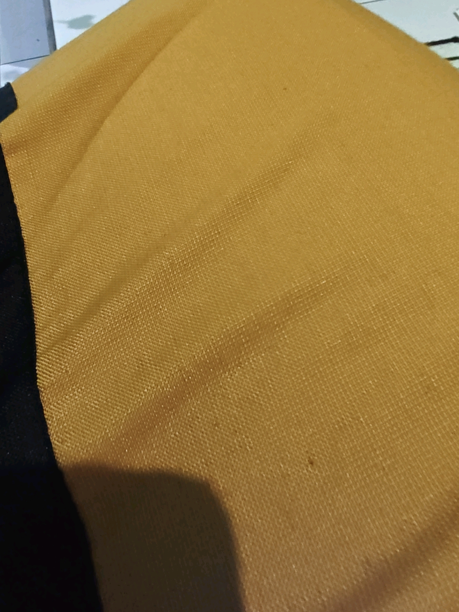 A close-up, high-angle shot shows the texture of a folded yellow fabric with a dark stripe on the left side. The fabric appears to be a synthetic material with a grid-like weave. The lighting casts a shadow on the lower right portion of the fabric. No location in Sumberwono, Indonesia is visible or implied.