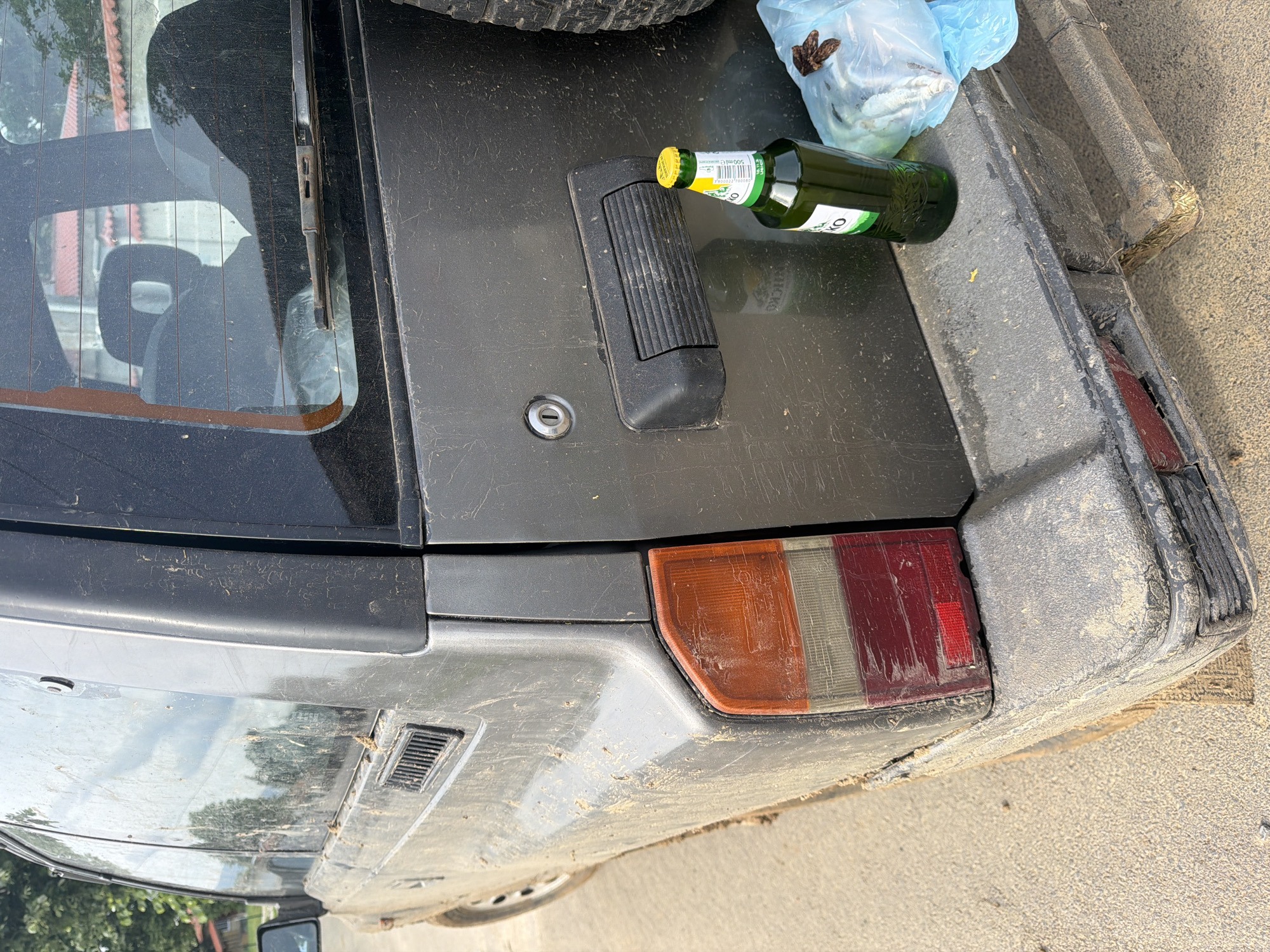The image captures the rear of a heavily dust-covered, dark gray or black car, likely an older model SUV or rugged hatchback, parked outdoors on a light brown, sandy or dirt surface. The vehicle's entire rear section, including the tailgate, rear window, bumper, and taillights, is coated in a thick layer of dry grime and dirt, suggesting extensive travel through unpaved or dusty terrain.
On the dirty tailgate, a green glass bottle with a bright yellow cap is prominently placed. The bottle's label displays "500 ml e" and a barcode; the brand name is partially visible, possibly containing Cyrillic characters. Adjacent to the bottle is a light blue plastic bag tied at the top, holding several small, pale objects that appear to be dead fish or similar aquatic creatures, alongside some dried, brown organic matter. This suggests activities such as fishing or foraging. A section of a black spare tire, indicative of a rugged vehicle, is visible in the upper left corner, possibly mounted on a roof rack or the rear door.
The rear window features horizontal defroster lines and a black wiper arm. A simple black handle/lock mechanism is centered on the tailgate. The car's right taillight, a rectangular unit with distinct red, amber, and clear sections, is visible but obscured by dirt and minor scratches. A small black vent is positioned on the lower left rear body panel. The bright daylight casts subtle shadows, and the overall scene conveys a rural or outdoor setting, consistent with the provided location context of Malomir, Bulgaria. No people are visible. The items on the car suggest a temporary stop after outdoor pursuits.