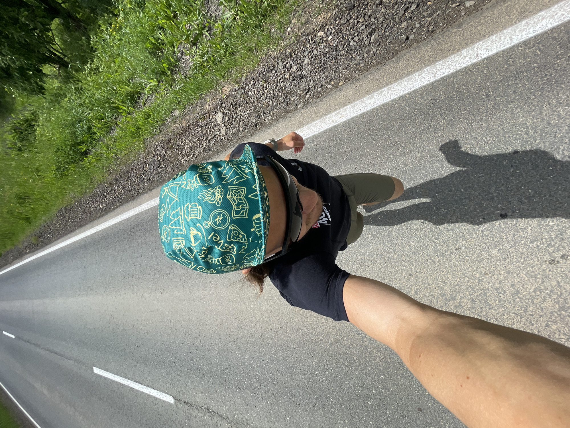 An overhead, tilted, first-person perspective shot captures a person moving along an asphalt road under bright daylight. The scene shows a person's upper body, right arm, and legs, along with a stretch of road bordered by green vegetation.
The person is wearing a teal-green cap adorned with numerous small white and yellow icons, including a pizza slice, a beer mug, a flame, a tree, an atomic symbol, and the word "PIZZA". They also wear sunglasses, a black t-shirt with partial white text visible (appearing to be "CALIBRATION" or similar), and olive green shorts. The person's right arm is extended, holding the camera, suggesting a selfie taken during an activity. A long shadow of the person is cast on the road to their right, indicating a high sun.
The road features a solid white line marking its edge on the right and dashed white lines in the center, extending into the distance. Lush green grass and dense foliage line the side of the road, suggesting a rural or natural setting. The overall impression is of an active outdoor pursuit on a sunny day.