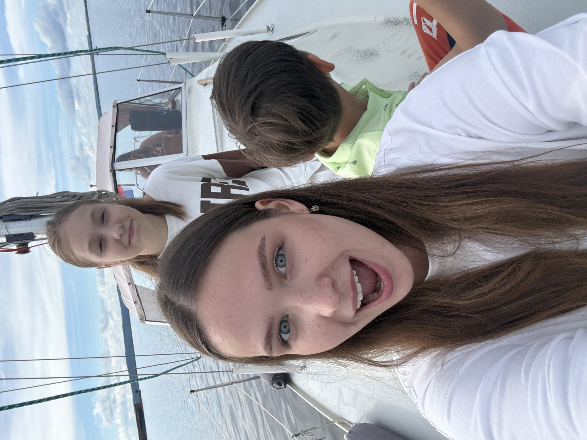 A group of three people are on a sailboat on a body of water, possibly the Kama River in Chaikovsky, Russia, given the surrounding landscape of water and distant tree-lined shores. In the foreground, a young woman with long brown hair and blue eyes is smiling widely at the camera, her mouth open. She is wearing a white t-shirt. Behind her and to the right, the back of a young boy's head is visible; he has short brown hair and is wearing a light green collared shirt. Further back, on the boat, another young woman with blonde hair tied back is looking towards the camera and smiling slightly. She is wearing a white t-shirt with brown lettering. A cabin structure is visible on the boat behind the two women. The sky is partly cloudy with blue patches.