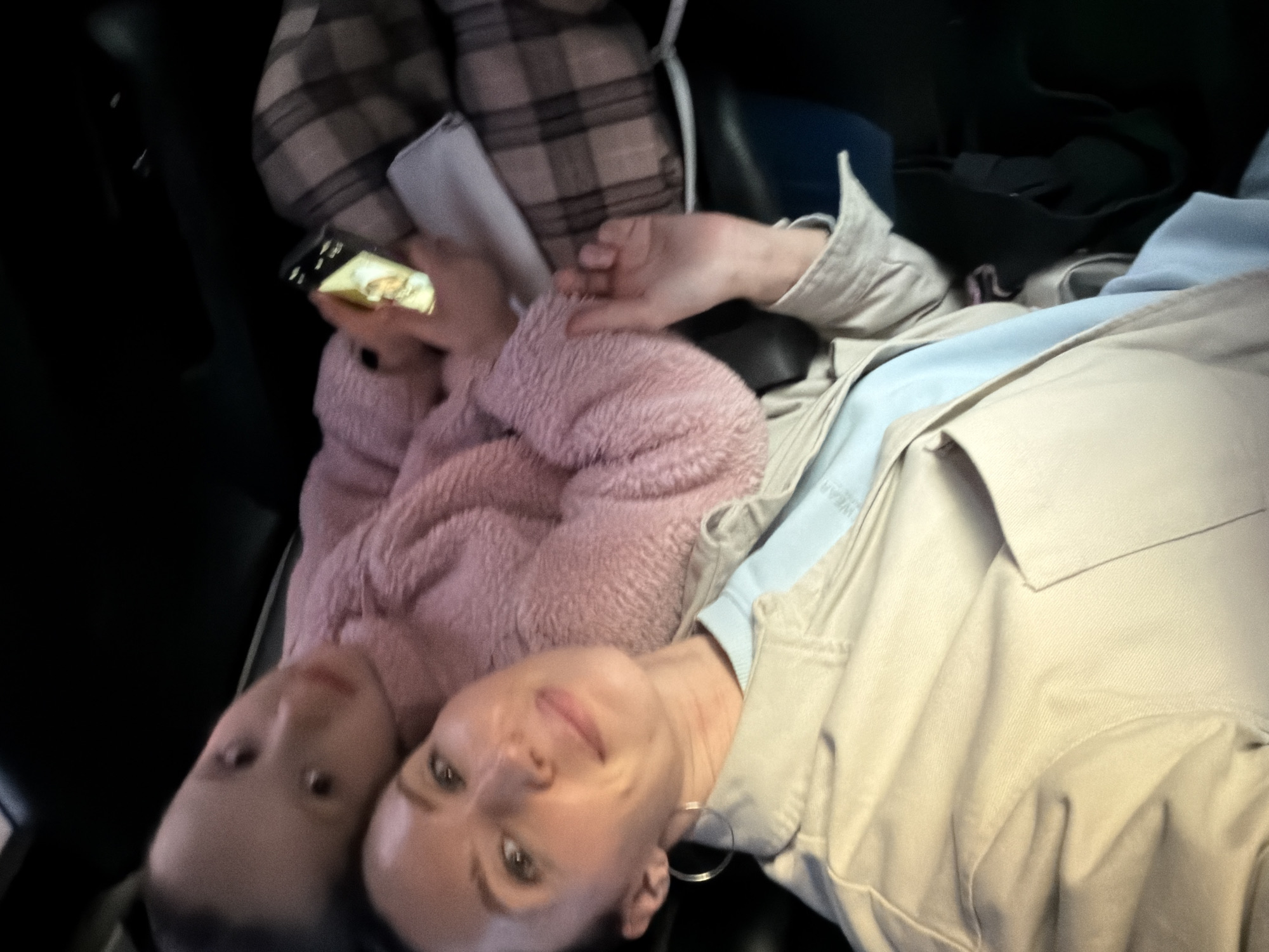 The image shows two individuals in what appears to be the interior of a vehicle, possibly a car or taxi. The lighting is dim, suggesting the photo was taken in the evening or inside a space with limited natural light. One person, a woman, is closer to the camera, looking directly at it with a slight smile. She wears a beige overshirt over a light blue top, and a hoop earring is visible. Another person, presumably a child, is next to her, wearing a fuzzy pink jacket. This person's gaze is directed to the left. One person holds a phone. There is a plaid bag and other objects in the background, contributing to a sense of being in a confined space. The environment is dark and cluttered, indicating a personal space such as a car interior. Without additional information, determining if this is in Perm, Russia is not possible from this image alone.