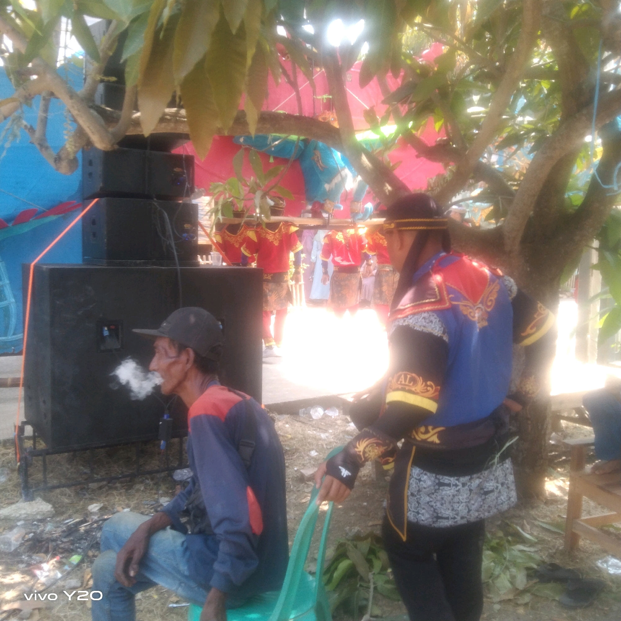 In an outdoor setting under a leafy tree, a man in a blue and red shirt and jeans sits smoking a cigarette, exhaling a cloud of smoke. He wears a dark baseball cap. To his right, two people are dressed in traditional, ornate costumes with red jackets and patterned skirts, and one has a decorative headband. Behind them, a large, bright light source illuminates the scene. Stacks of black speakers are visible to the left, and the ground is covered in dry leaves and debris. In the background, colorful fabric, possibly a tent or canopy, is visible. The overall impression is of an outdoor event or performance.