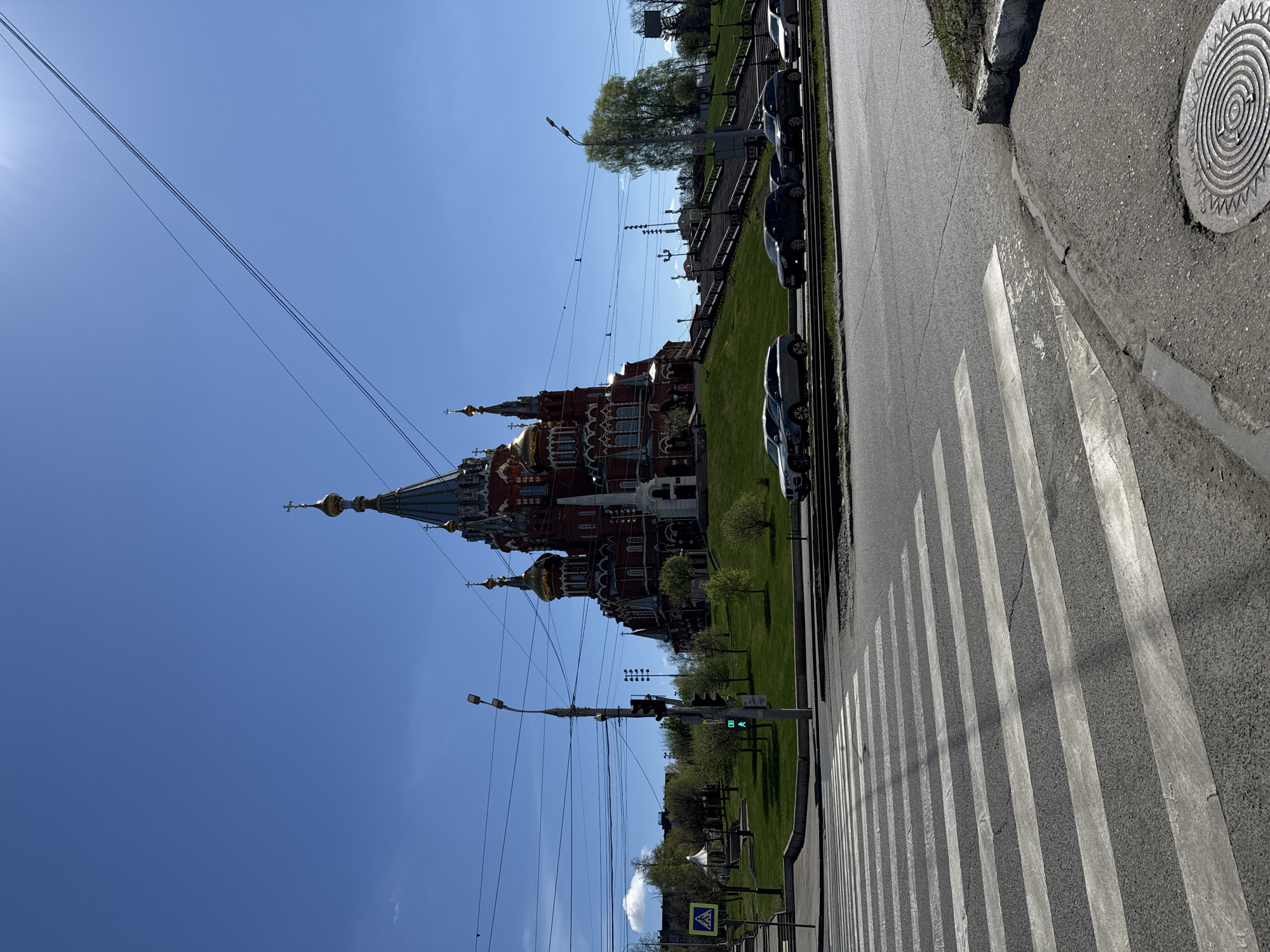 The image depicts the Cathedral of Alexander Nevsky in Izhevsk, Russia, viewed from across a street. The cathedral is a large, red-and-white building with gold domes, situated on a green lawn. Along the roadside, several cars are parked parallel to the curb. A pedestrian crosswalk stretches across the foreground of the street. Overhead wires crisscross the sky above the cathedral. A traffic light displays a green signal. The sky is clear and blue.