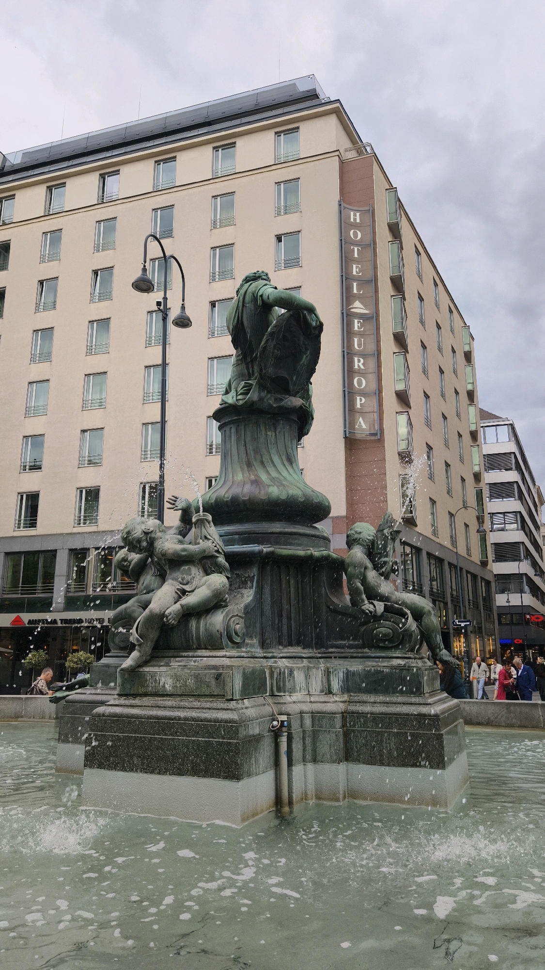The image depicts the Donnerbrunnen fountain in Vienna, Austria, in front of the Hotel Europa. The fountain is a large, ornate bronze sculpture featuring several cherubic figures surrounding a central column. Water is spraying from various points of the fountain, creating a dynamic effect. In the background, the Hotel Europa is a multi-story building with a prominent vertical sign spelling out "HOTEL EUROPA." The sky is overcast, suggesting a cloudy day. Several people are visible in the background on a sidewalk, some walking and others standing. One person is seen sitting on the edge of the fountain in the lower left portion of the frame. The water in the fountain's basin is a murky green color.