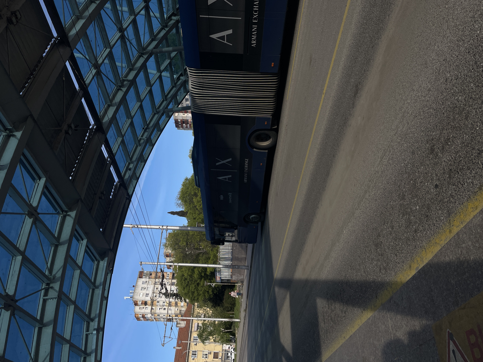 The image shows an articulated bus with "A|X ARMANI EXCHANGE" branding parked at a bus stop in Burgas, Bulgaria. The bus stop has a modern glass and metal roof structure. A pedestrian is on the sidewalk near the bus stop. Buildings, trees, and overhead power lines are in the background. The ground is asphalt with yellow lane markings. The sky is blue.