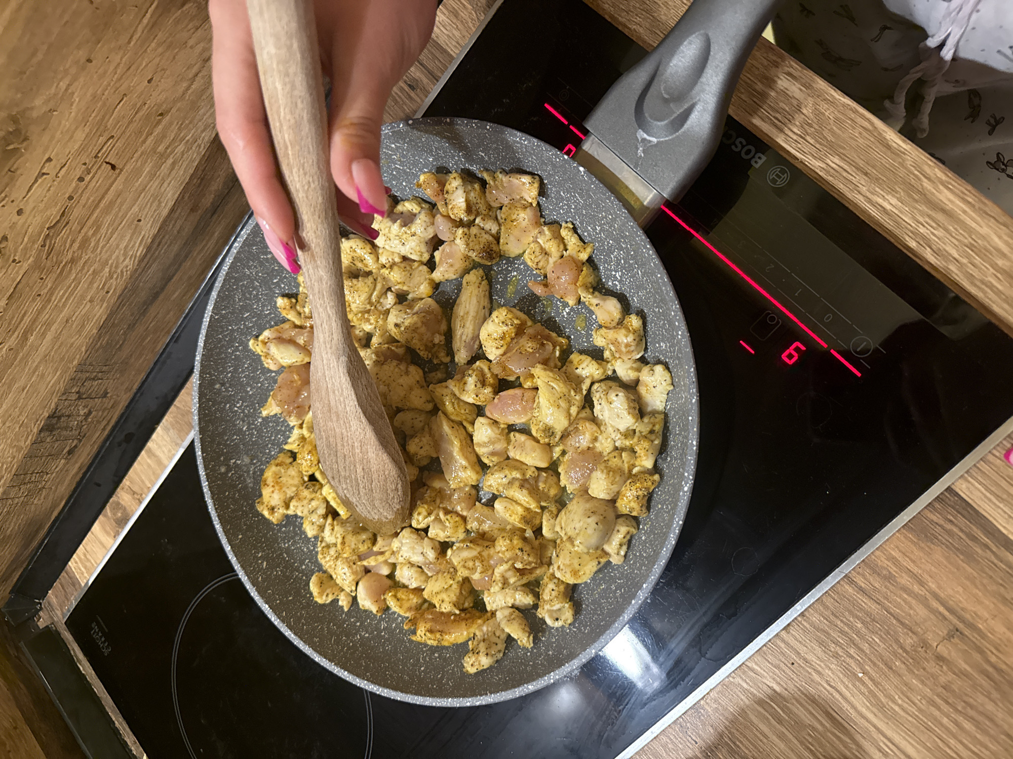 An overhead, medium close-up shot captures a person cooking chicken in a kitchen setting. The central focus is a gray non-stick frying pan, brimming with numerous small, seasoned chicken pieces, resting on a black induction cooktop. A person's right hand, distinguished by long, almond-shaped nails with a vibrant pink French manicure, holds a light-colored wooden spoon, positioned over the chicken, suggesting an action of stirring or mixing.
The chicken pieces are irregularly cut cubes, coated in a yellowish seasoning with visible dark specks of pepper or spices. Some pieces still show a pinkish hue, indicating they are raw or partially cooked. The induction cooktop is active, displaying glowing red lines that delineate an energized cooking zone. A red LED display on the cooktop clearly shows the number "6," likely indicating the current heat level. The brand name "Bosch" is prominently visible on the cooktop's surface. Faint text, "SCOTT CERAN," a type of glass-ceramic, can also be discerned on the far left burner.
The cooktop is integrated into a warm, brown wooden countertop, which features a distinct grain pattern and frames the cooking area on both sides. In the upper right corner of the background, a glimpse of a light-colored fabric with a subtle, dark pattern (possibly animals like bunnies or deer) is visible, potentially part of an apron or clothing. The scene depicts a domestic kitchen, likely indoors in Varna, Bulgaria. No specific time of day, weather conditions, or emotions are discernible. The activity is clearly the preparation of food, specifically sautéing or frying chicken.