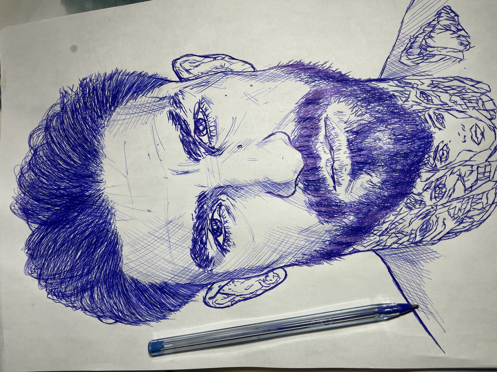 A close-up, eye-level shot captures a detailed pen-and-ink portrait of a man drawn on white paper. The portrait is rendered in blue ink, with visible cross-hatching and linear strokes used to create shading and texture. The man has short, dark, and somewhat messy hair, a beard, and a mustache. His eyes are looking directly at the viewer. There are tattoos visible on his neck, featuring multiple faces and intricate designs. To the left of the portrait, a clear plastic ballpoint pen with a blue ink tip is positioned vertically, with its tip resting on the paper. The background is a plain white surface. There is no indication of a specific location in Jos, Nigeria, within the image.