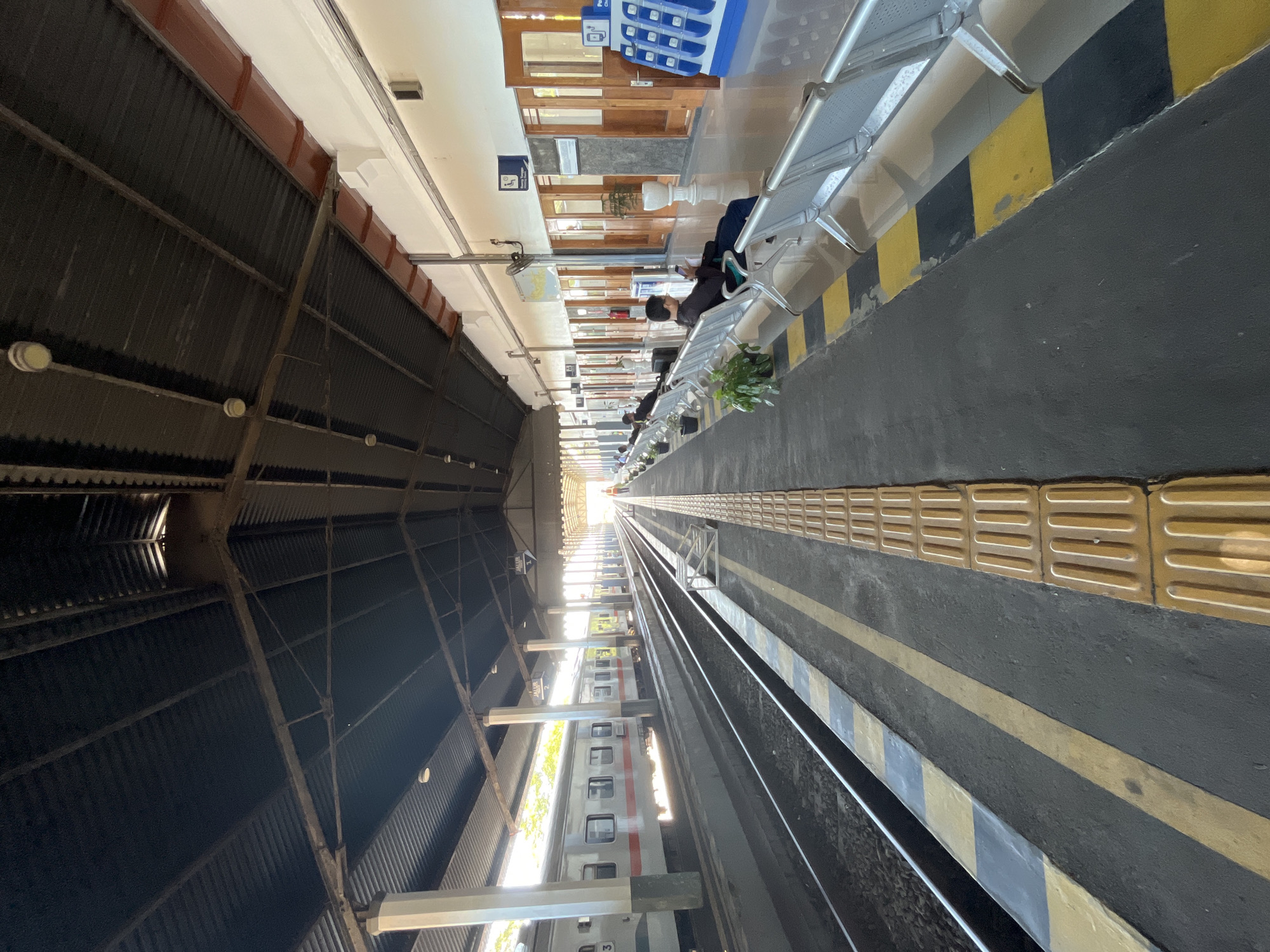 The image depicts a train station platform in Desa Tambakreja, Indonesia. The platform is covered by a dark corrugated metal roof supported by white pillars. Two train tracks run parallel along the platform. On the left side, a white and red train is stopped at the platform. On the right side, several rows of empty silver benches are arranged for waiting passengers. Some potted plants are placed near the benches.
Further down the platform, a few people are seated on the benches, some looking at their phones. A charging station with multiple ports is visible on the right side of the platform. The walls of the station building are painted white and feature several wooden doors and windows. Signs are mounted on the wall, including one with an "i" symbol and text that appears to be in Indonesian. The ground of the platform is paved with dark asphalt, marked with yellow and black lines for safety. A tactile paving strip is present along the edge of the platform. Sunlight streams in from the end of the platform, creating a bright, elongated perspective.