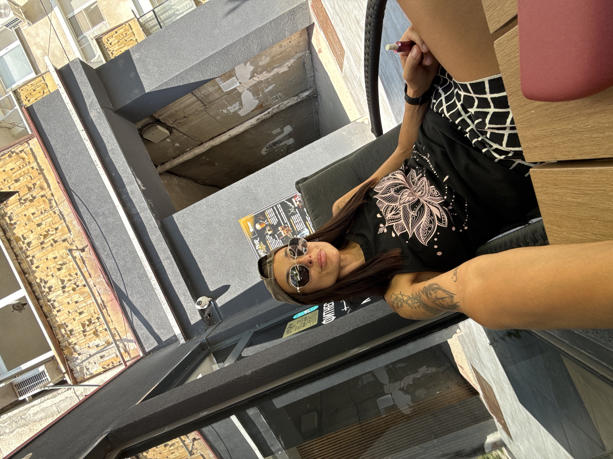 A woman is sitting in a chair outside a building, holding a vape. She is wearing sunglasses, a baseball cap, a black tank top with a pink floral design, and black and white checkered shorts. Tattoos are visible on her arm. The background shows a building with a textured yellow facade, a grey painted exterior, and a narrow entryway. The scene appears to be in the city of Pleven, Bulgaria, based on the Cyrillic text on a sign visible in the background. The overall impression is a casual outdoor setting on a sunny day.