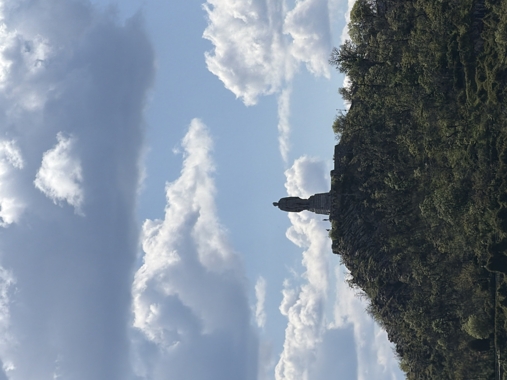 The image presents a majestic stone statue positioned high atop a rugged, tree-covered hill under a partly cloudy sky. The statue, a tall, upright figure, likely male, stands on a prominent, multi-tiered pedestal, appearing as a significant historical monument. It is silhouetted against the sky, with some architectural details of its base and form discernible.
The hill supporting the monument is densely covered in various shades of green foliage and trees, interspersed with visible rocky textures and outcrops, suggesting a natural and wild landscape.
The sky above features a blend of bright, fluffy white cumulus clouds and more expansive, muted grey cloud formations, with areas of clear blue sky peeking through. The lighting indicates it is daytime, with natural illumination across the scene.
No specific activity or event is taking place; the image captures a static view of the monument within its natural setting. There are no clearly identifiable people or any discernible text visible in the frame. The scene conveys a sense of grandeur and historical presence integrated with the natural environment.