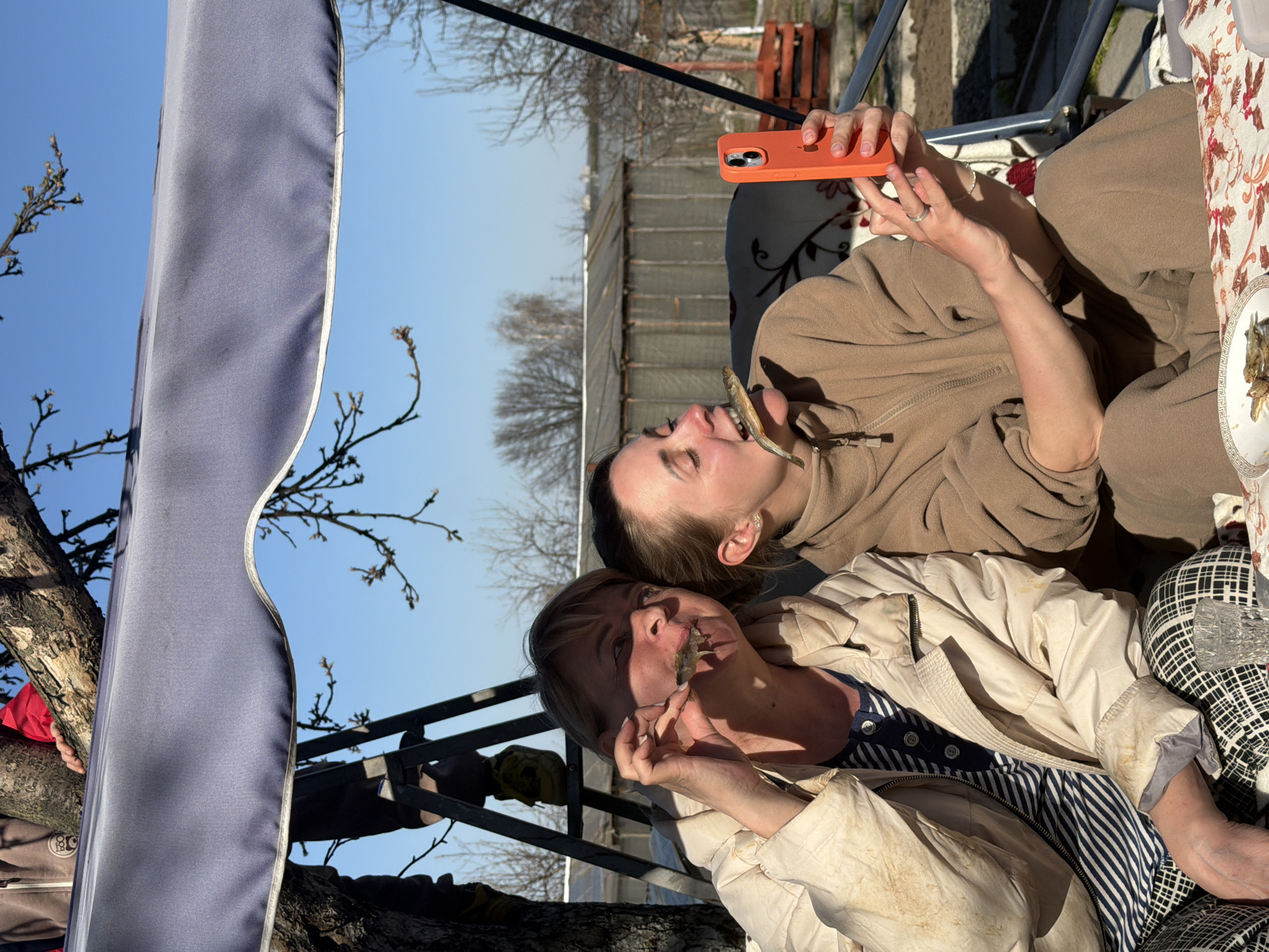 Two women are seated outdoors and appear to be taking a photo. The woman closest to the camera is wearing a tan jacket, a blue and white striped shirt, and holding a small fish to her mouth. The other woman is wearing a tan fleece, holding an orange smartphone, and has a small fish in her mouth. They appear to be sitting under a canopy or awning attached to a tree. The background includes a fence, trees without leaves, and a clear blue sky. The location is Chaikovsky, Russia.