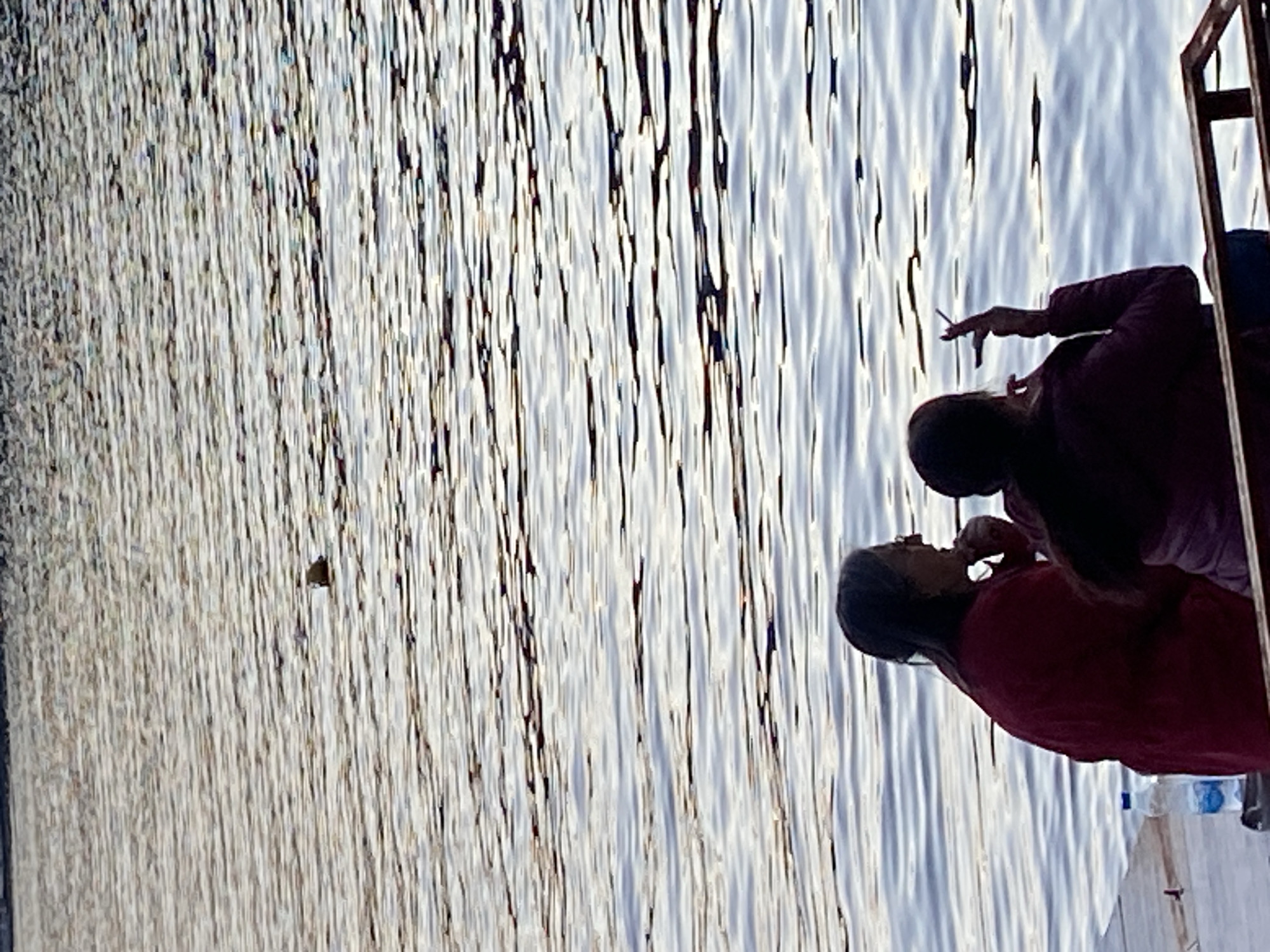 Two people are on a wooden structure over the water. One person is smoking a cigarette. The water is rippled with reflections of the sun. Plastic water bottles are visible on the edge of the structure. The location is likely a body of water in or around Budva, Montenegro.