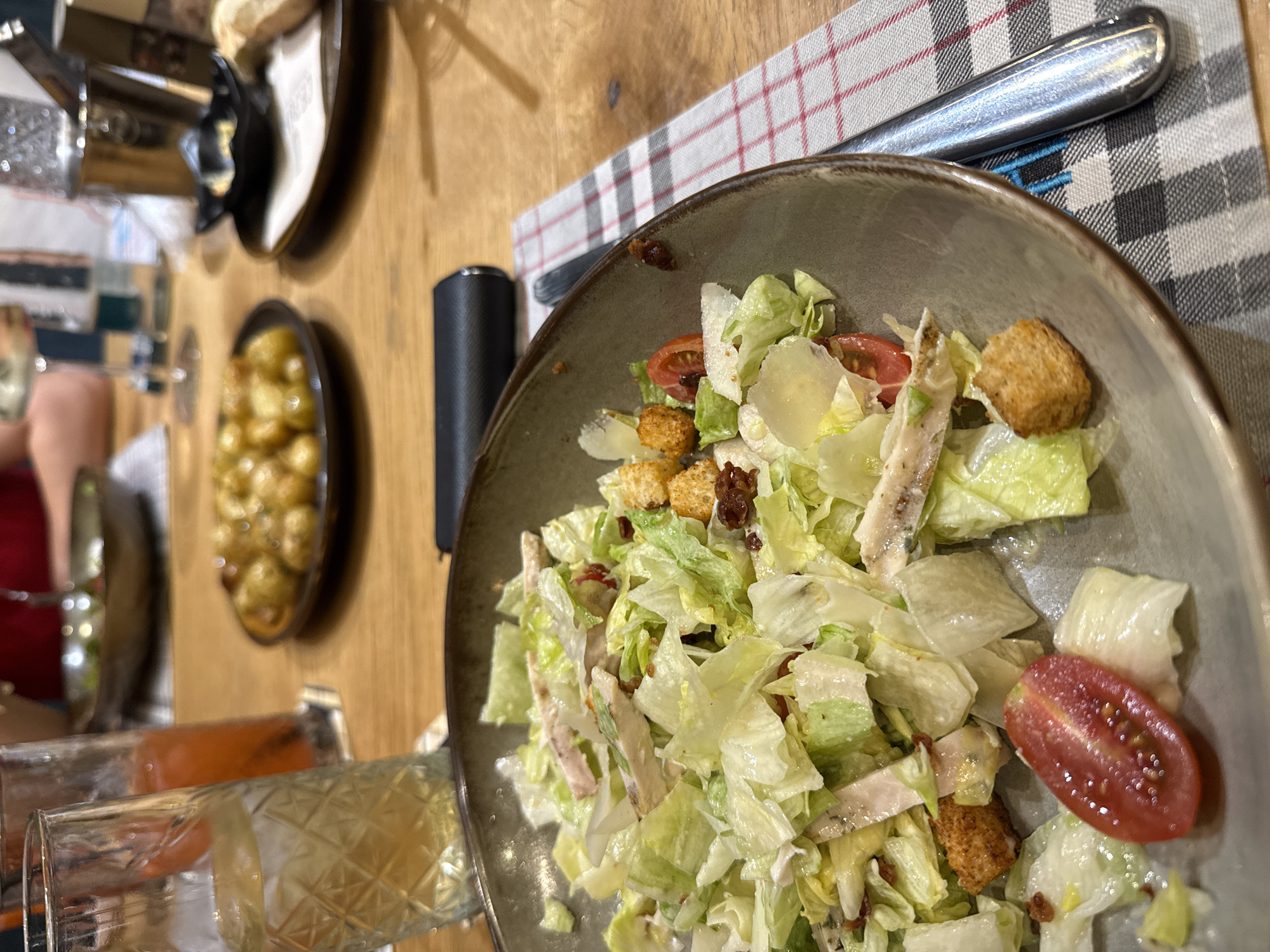 The image presents an overhead view of a dining table, focusing on a large bowl of salad. The salad, which appears to be a Caesar salad, is comprised of crisp chopped lettuce, sliced chicken pieces, golden croutons, bright red cherry tomato halves, and thin shavings of what looks like Parmesan cheese, all coated in a light dressing.
The salad bowl rests on a grey and white checkered placemat with subtle red criss-cross lines. A silver fork is positioned on the placemat to the right of the bowl. To the immediate left of the salad bowl, a black, cylindrical object, possibly a salt and pepper grinder or a similar dining accessory, is visible.
The dining table is made of light-colored wood. In the blurred background, other elements of a meal are discernible. To the left, a small, dark oval dish holds numerous round, golden-brown items, possibly roasted potatoes or gnocchi. Further towards the foreground on the left, two glasses are present; one contains a light, clear or yellowish beverage, and the other holds a reddish or orange-colored drink. Faint blurs of people, including arms and clothing, suggest that this is a shared meal or gathering. The scene conveys a casual and relaxed atmosphere, likely an indoor setting with warm lighting, potentially during an evening meal in Sofia, Bulgaria. No explicit text is visible.