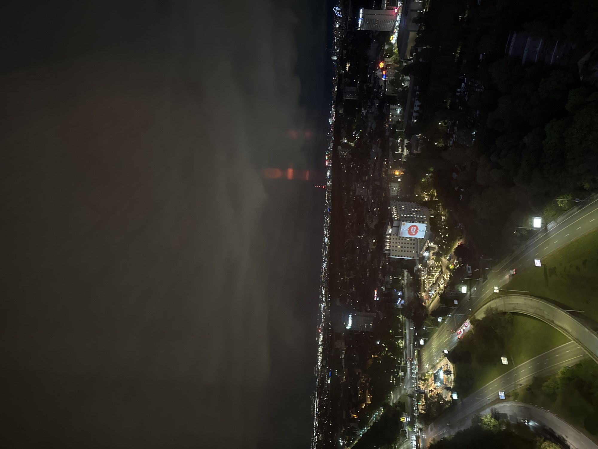 The image is a nighttime aerial view of Sofia, Bulgaria. To the right of the image, a lit road, likely a highway, curves through a green, wooded area. A building with a large advertisement is visible amidst the city's lights. In the distance, along the horizon, the city lights blur into an illuminated line. To the left, the image shows an area covered with dense fog or clouds, reflecting the city lights. The city's density appears to increase towards the center of the image.