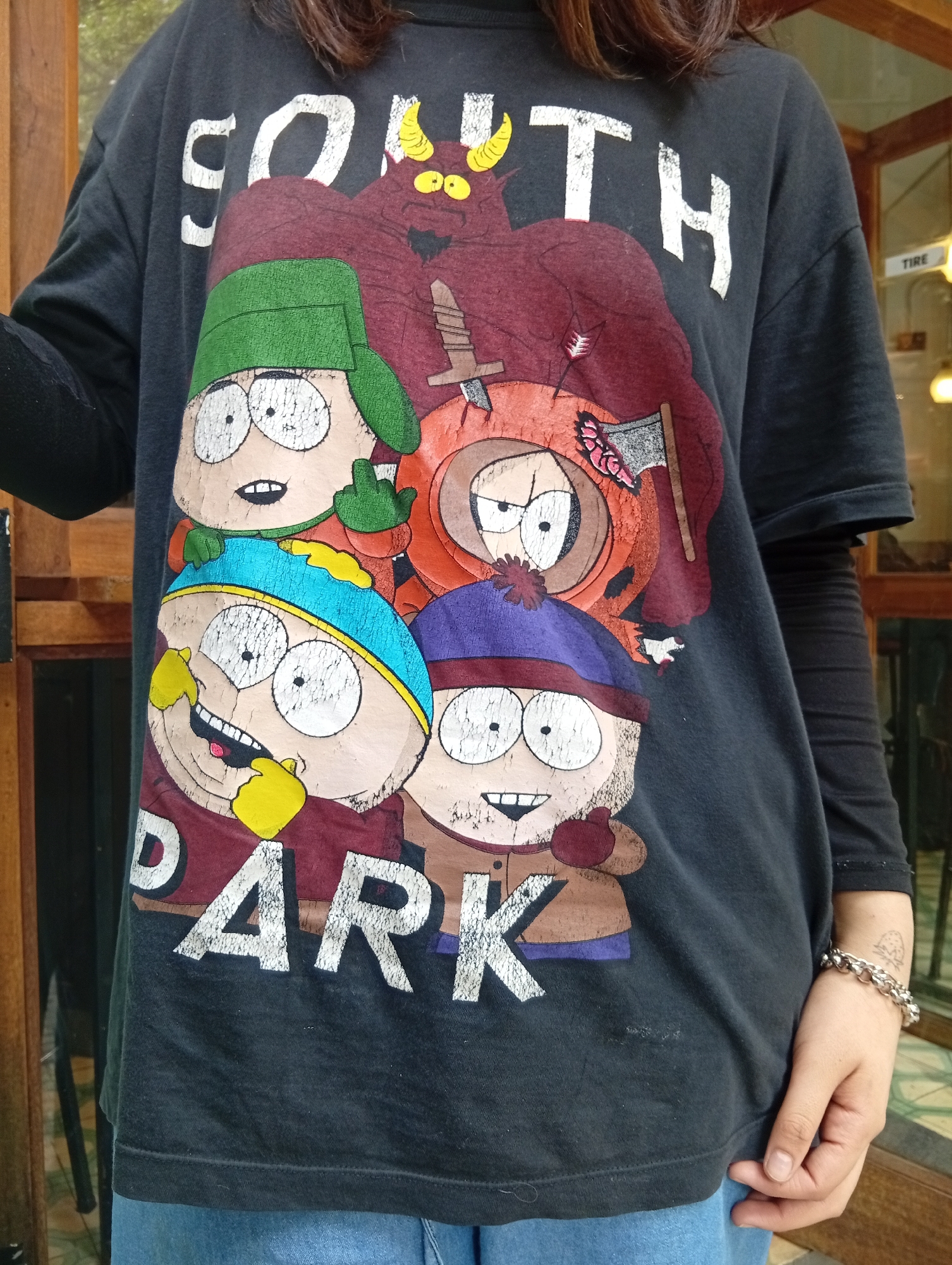 A person wearing a black "South Park" t-shirt is standing indoors. The t-shirt graphic includes the characters Cartman, Kyle, Stan, Kenny, and Satan from the animated TV show "South Park". The person wears a long-sleeved black undershirt and blue jeans. The person is wearing a silver chain bracelet on their left wrist.