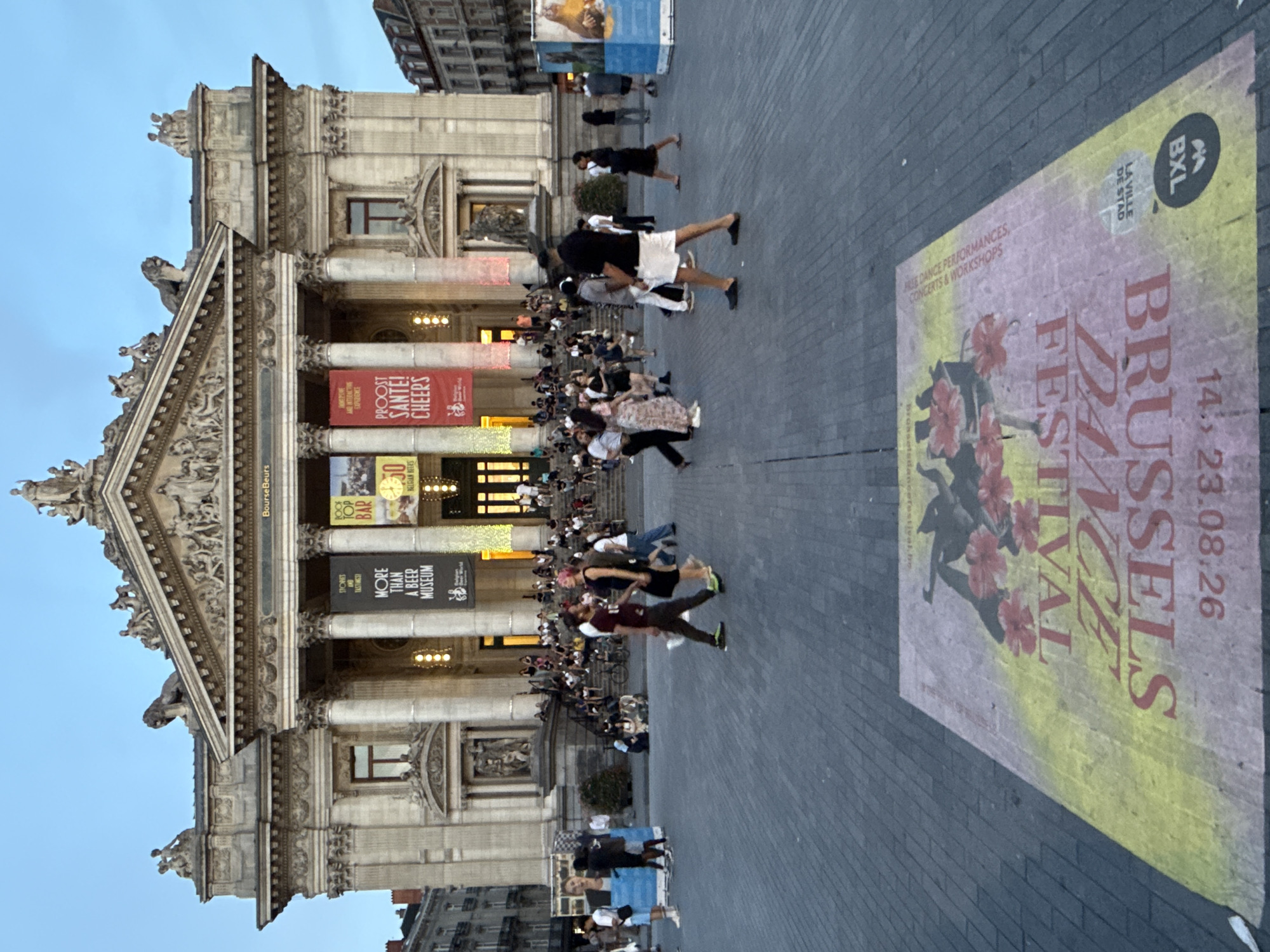 The image captures a bustling evening scene in Brussels, Belgium, specifically in front of the Brussels Stock Exchange Building. The grand neoclassical facade of the building dominates the background, adorned with ornate sculptures and columns. Several large banners hang from the building, promoting events, including one for "More Than A Beer Museum" and another that reads "PROOST! SANTE! CHEERS!".
The foreground is a wide, paved square where numerous people are gathered and walking. The paving stones are laid in a patterned arrangement. A large, colorful advertisement is laid flat on the ground in the lower portion of the image, featuring text in red that reads "BRUSSELS DANCE FESTIVAL" and a date which appears to be "14. 23.08.26". The advertisement also includes imagery of flowers.
People of diverse ages and ethnicities are present, dressed in casual attire. Some are walking across the square, while others are congregated in groups, possibly socializing or observing. A few individuals are seated on the steps leading up to the building's entrance. The lighting suggests it is either dusk or early evening, with artificial lights illuminating parts of the building and the surrounding area. In the distance, other city buildings are visible, indicating an urban environment. The overall atmosphere is one of public gathering and activity.