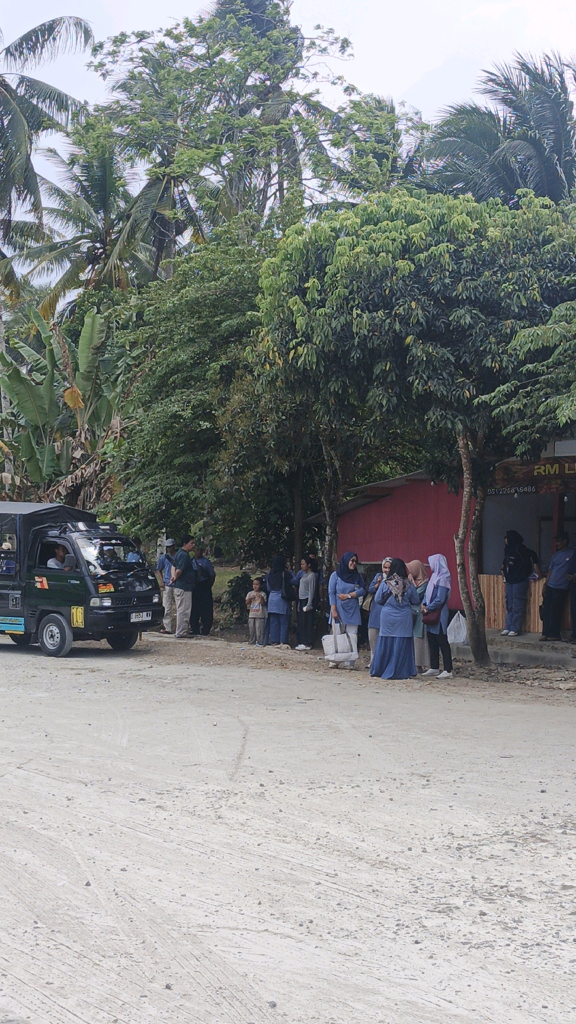 The image shows a roadside scene in Cibenda, Indonesia, with several people standing near a small red building and a green truck. The building appears to be a shop or a small establishment, with a sign that reads "RM LU". A phone number, "051226838486", is also visible on the building. The truck, a utility vehicle with an open bed, is parked on the left side of the image.
Several individuals are gathered on the sidewalk in front of the building, some of whom are wearing headscarves. One person appears to be driving the truck. The environment is characterized by lush green trees and vegetation, indicating a rural or semi-rural setting. The ground is unpaved, consisting of dirt and gravel. The sky is overcast.