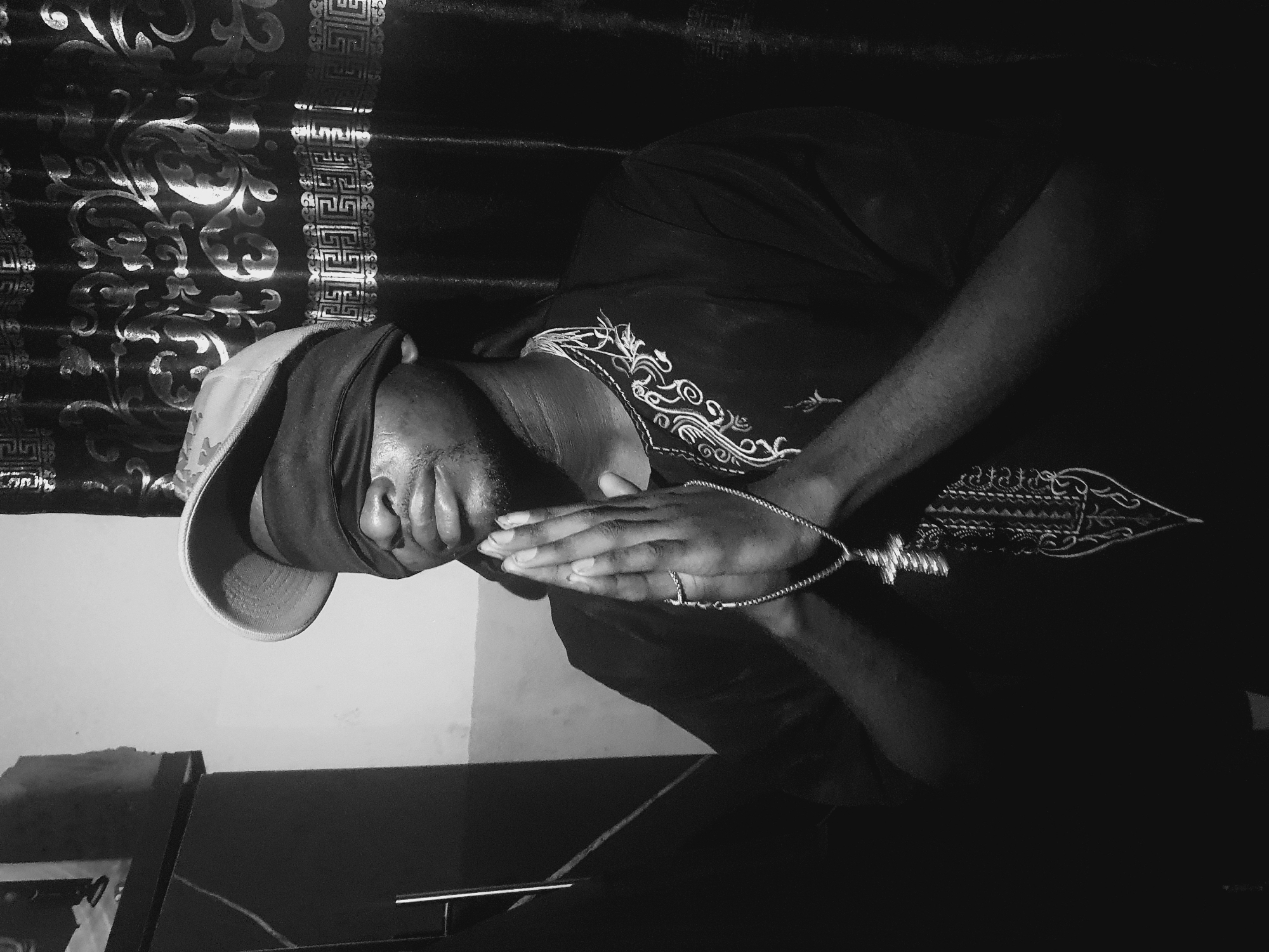 A man wearing a cap and patterned shirt clasps his hands in front of him, possibly in prayer. He is also wearing a necklace with a decorative pendant. The scene is indoors with patterned curtains in the background. The location is unknown.