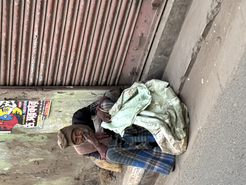 A homeless person sits wrapped in blankets and clothing under a corrugated metal awning. They are positioned against a concrete wall next to a poster. The individual wears a knit cap and a patterned scarf around their neck. A light-colored blanket partially covers them. The ground is rough and weathered, suggesting an outdoor setting.