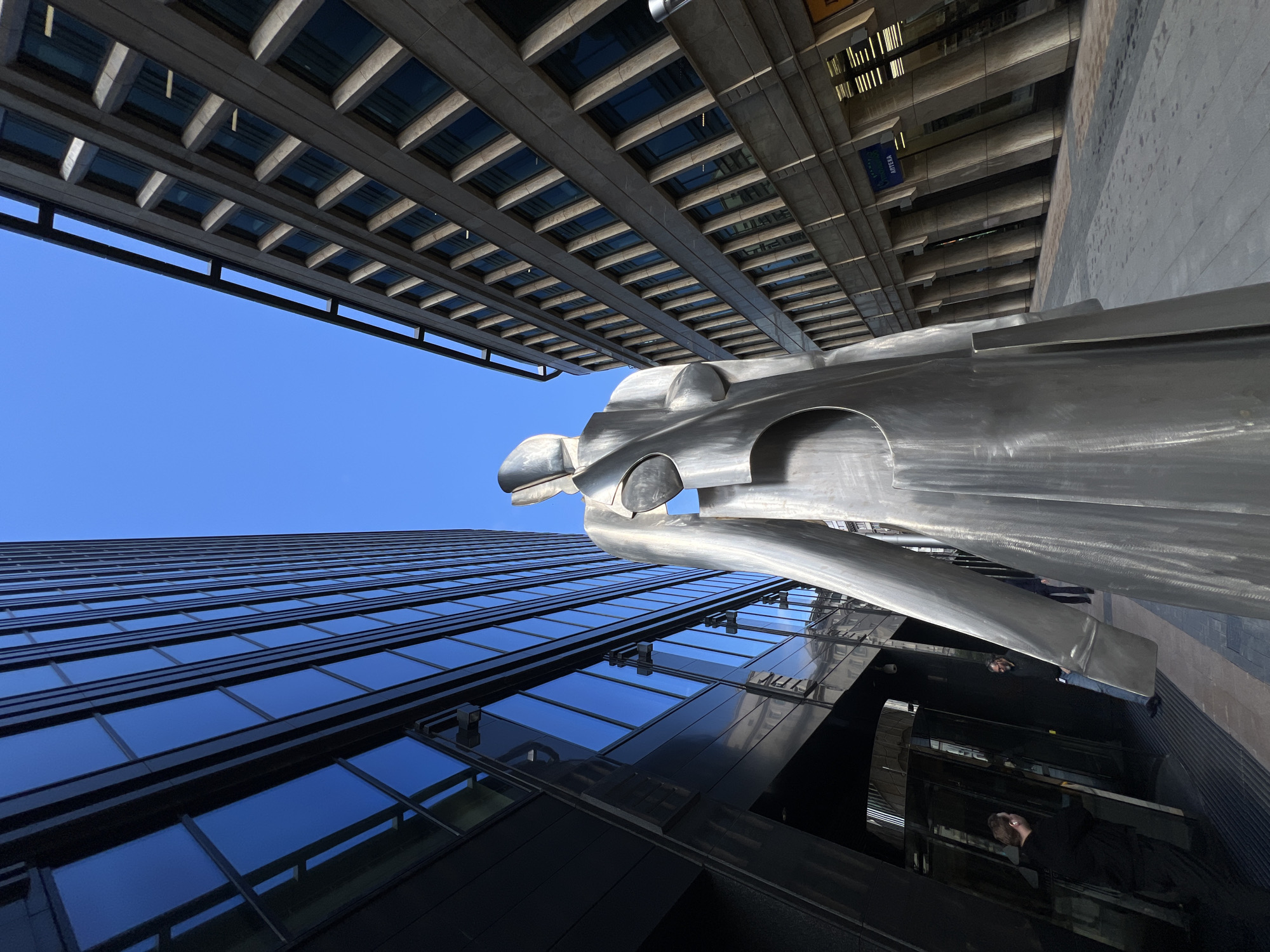 The image, taken from a low angle, features a metallic sculpture of a stylized figure suspended between two tall buildings in Moscow, Russia. The building on the left has a façade composed of dark glass windows set within black frames. The building on the right exhibits a concrete structure with recessed windows. The sky above is clear and blue. The sculpture itself appears polished and reflective. The image suggests an urban environment with a focus on modern architecture and public art. Reflections on the building reveal people walking.