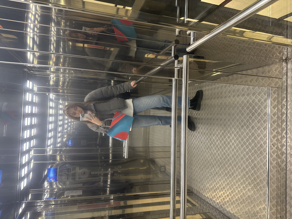 A young adult female is depicted standing centrally inside a modern, highly reflective elevator, actively taking a mirror selfie with a silver smartphone held in her raised right hand. She is dressed in a grey cropped puffer jacket over a white t-shirt, light blue jeans, and black platform boots. In her left hand, she holds a shopping bag with a distinctive geometric pattern in red, light blue, and white. The elevator interior features polished metal walls that reflect the subject and the internal environment, including square recessed overhead lighting. Two horizontal polished metal handrails traverse the lower half of the frame. The floor is composed of textured metal with a raised diamond plate pattern. On the left wall of the elevator, a blue digital display clearly shows the number "2" and the text "МОНТЭН ПАРК", identifying the location as an elevator within the Monten Park residential complex, specifically at Usacheva St, 11/1, Moscow, Russia.
