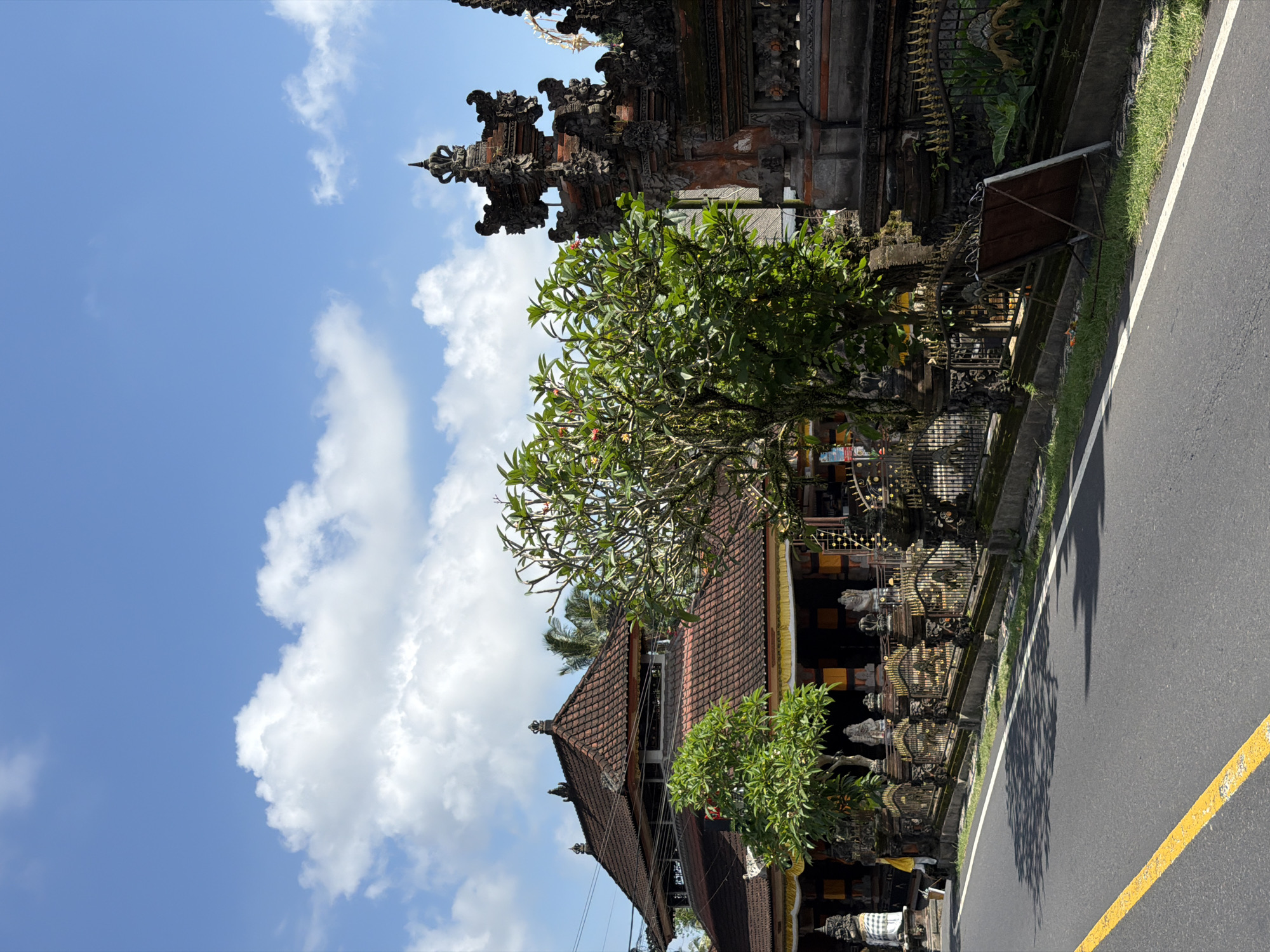 The image depicts a street scene in Tampaksiring, Indonesia, on a bright, sunny day. The sky is a vibrant blue with fluffy white clouds. On the left side of the frame, a traditional Balinese temple complex is visible, characterized by intricate stone carvings and ornate architecture. A tree with lush green leaves and some red flowers stands in front of the temple, partially obscuring some of its details. The temple features multiple tiers of roofs and decorative elements common in Balinese religious structures.
To the right of the temple is a paved road with a yellow line marking the edge. The road appears to be well-maintained. There is a strip of green grass and a low stone wall separating the temple from the road. The overall atmosphere suggests a calm and serene environment, typical of a rural or semi-rural setting in Bali. No people are clearly visible, and the focus is on the architectural elements of the temple and the natural surroundings. The time of day appears to be mid-morning or early afternoon, given the strong sunlight casting shadows.