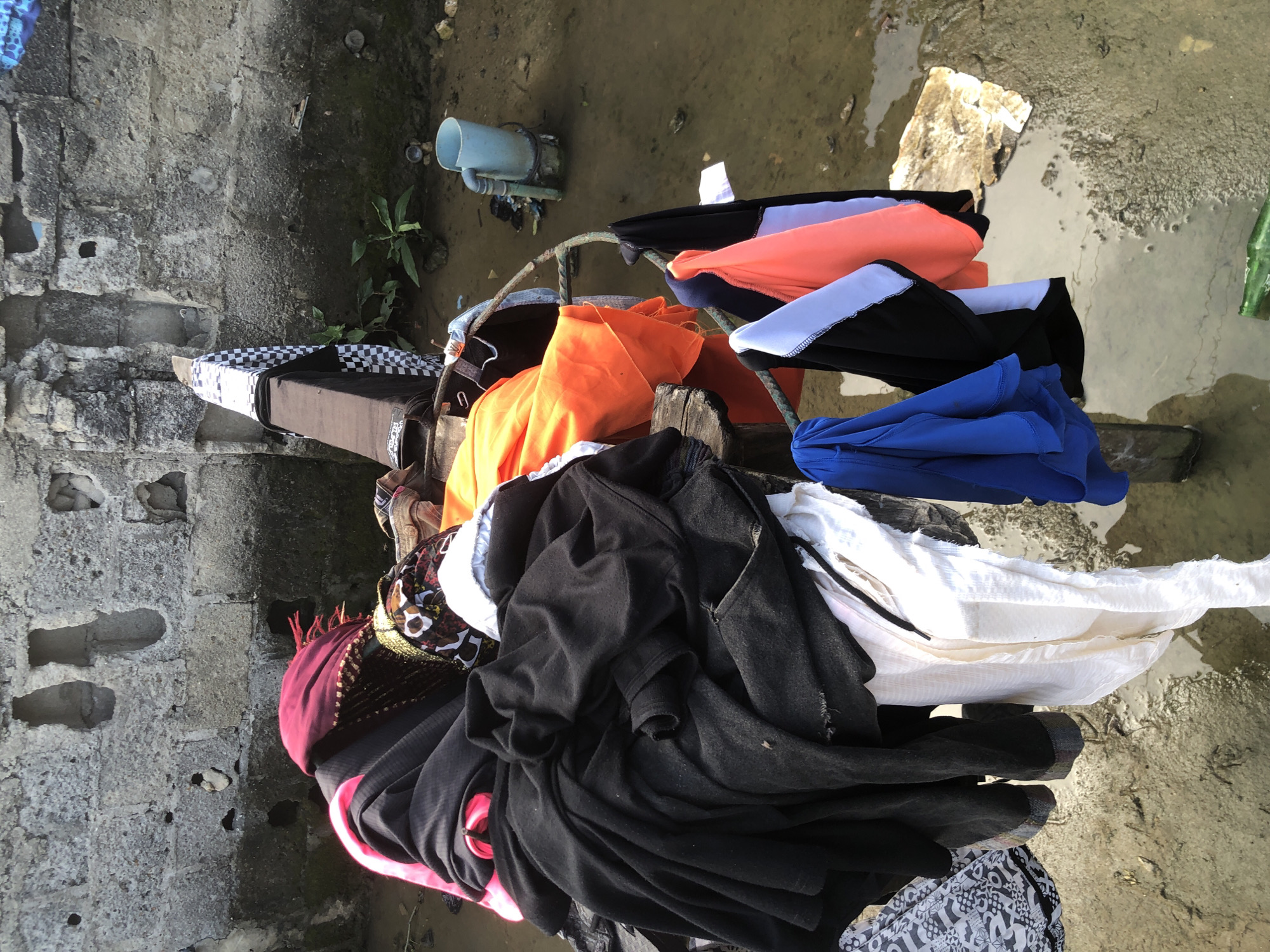 In Ado, Nigeria, a collection of laundry is draped over a makeshift drying rack constructed from wood and metal. The rack is positioned over a muddy, shallow body of water, possibly a drainage ditch or a flooded area. The laundry consists of various garments, including black, white, orange, and blue clothing, some of which appear to be folded or piled high. In the background, a rough concrete wall with visible holes and a patch of green vegetation can be seen. A light blue plastic container with a handle is also present near the water's edge. The overall scene suggests an outdoor setting where laundry is being air-dried in less than ideal conditions.