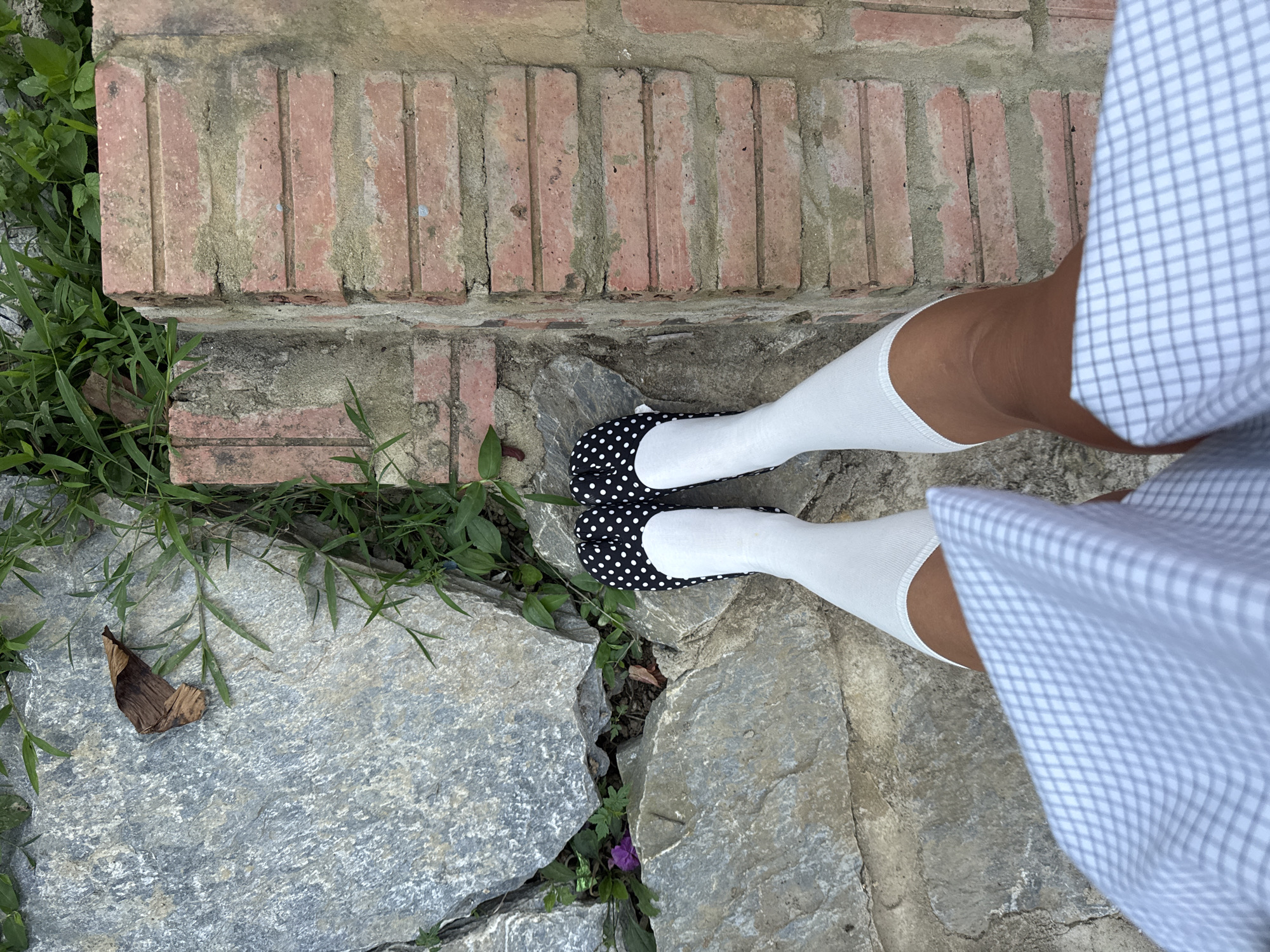 The image shows a person standing on a stone path next to a brick border, presumably outdoors. The person is wearing white knee-high socks and black and white polka-dot shoes. A light blue and white checkered dress is visible at the edge of the frame. A dry leaf lies on the stone path near some greenery. The path is cracked, and some greenery is growing in the cracks. The exact location in Bắc Hà, Vietnam, is not discernible from the given image.