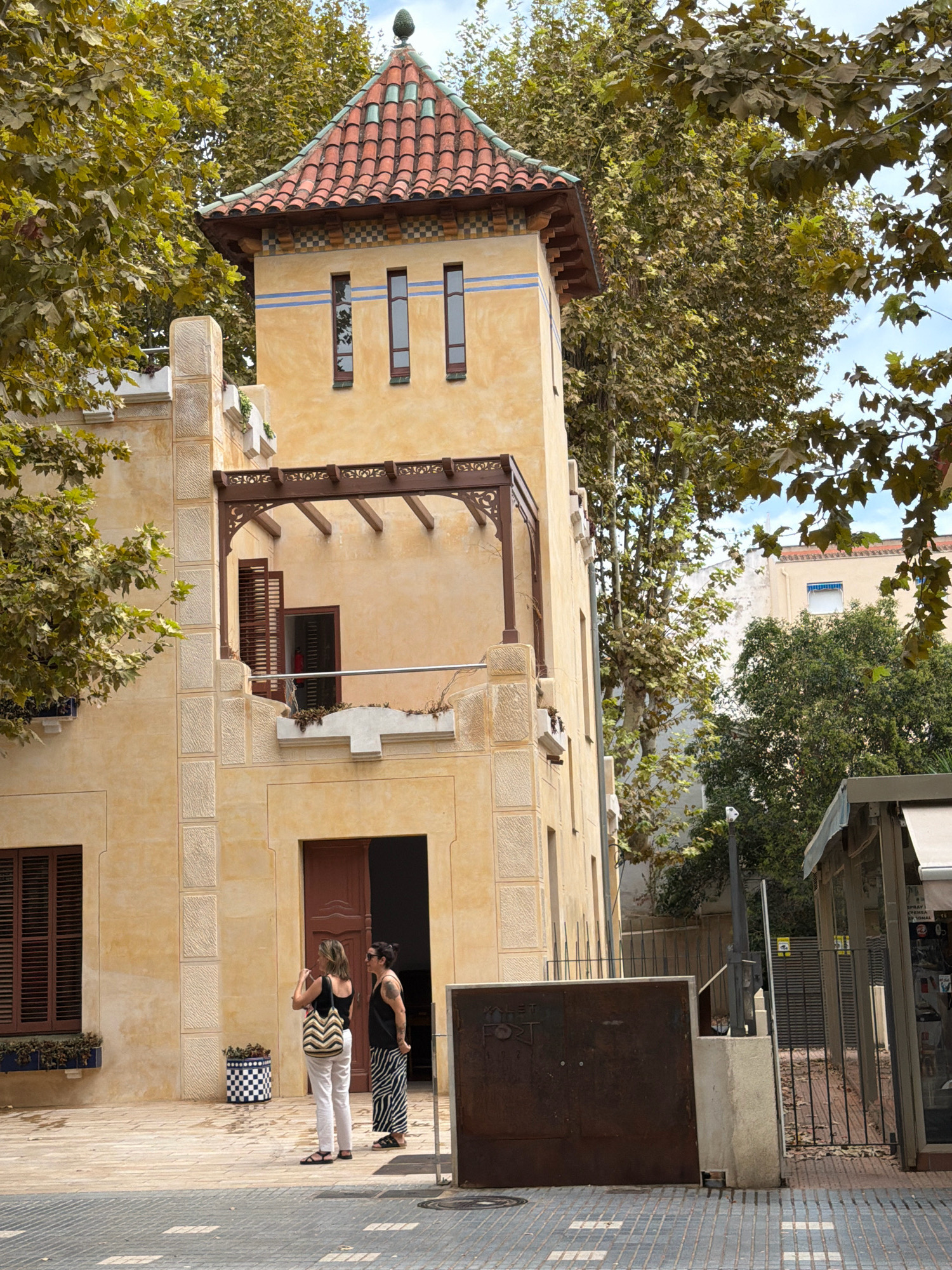 The image shows a two-story building with a distinctive red-tiled, multi-tiered roof and a central tower. The building's facade is a pale yellow with textured square insets and decorative blue stripes near the top of the tower. A wooden balcony with ornate carvings extends from the second story.
In front of the building, two women are standing on a paved patio. One woman, wearing white pants and a black top, holds a bag with a chevron pattern. The other woman, wearing a black and white striped skirt and a black top, has tattoos on her arms. They appear to be interacting casually, with the woman in white looking towards the building and the woman in stripes looking down.
To the right of the building, there is a black metal fence and a structure with a white awning, possibly a kiosk or a shop. Trees with green and yellowing leaves surround the building and extend into the background, with a blue sky visible above. The paving stones in the foreground are laid in a pattern of grey and white squares.
This building is identified as "Casa Bonet," located in the city of Salou, Spain, on Carrer del Progrés. It is a modernist building designed by architect Domènec Sugrañes i Gras and built between 1915 and 1918.