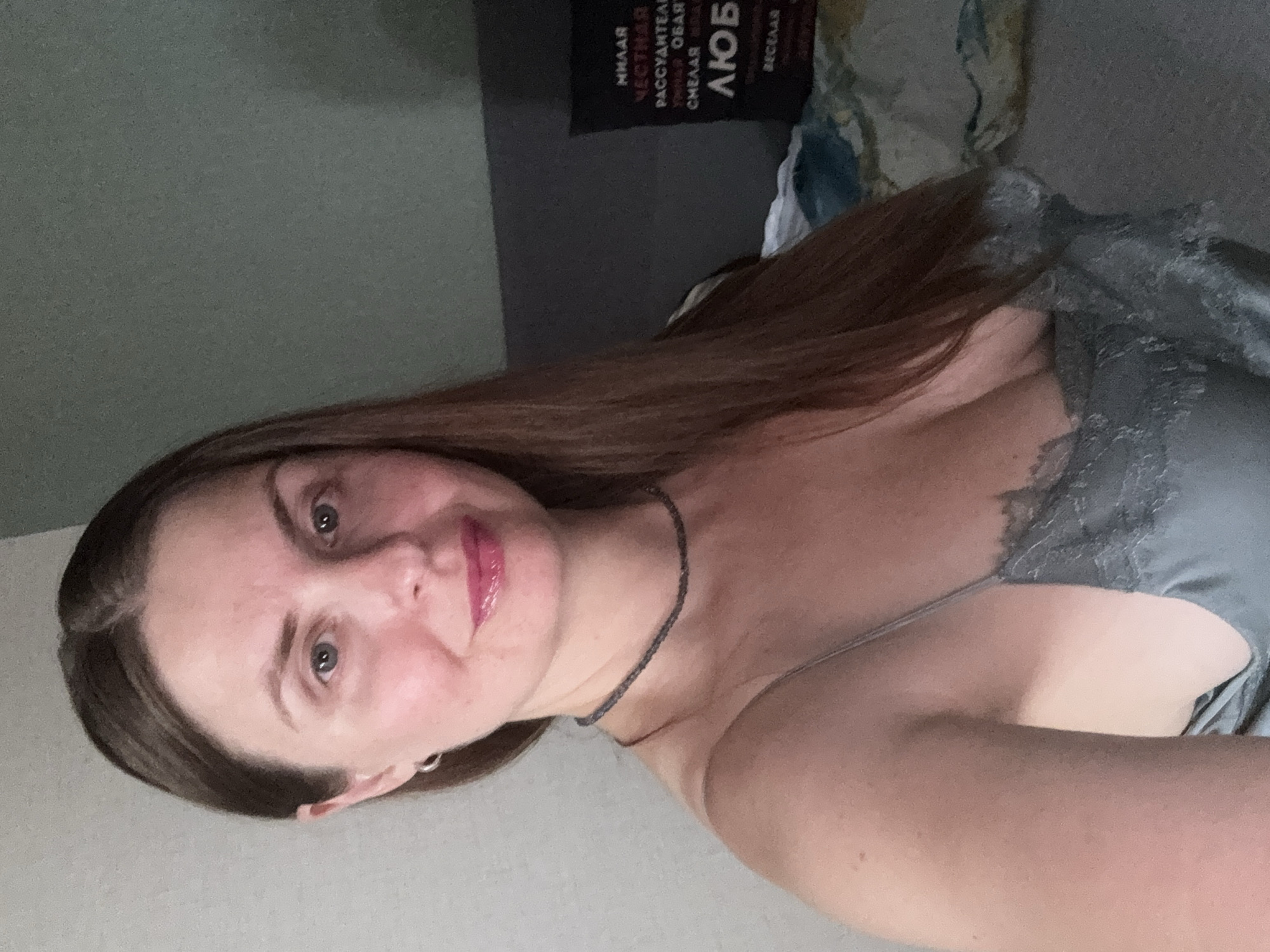 A woman with long, brown hair and blue eyes is looking at the camera. She is wearing a gray slip with lace trim and a thin necklace. The background is a wall and a bed with some pillows. There is text on one of the pillows that is illegible. The image appears to have been taken indoors, possibly in a bedroom. The location is the city of Konakovo, Russia.