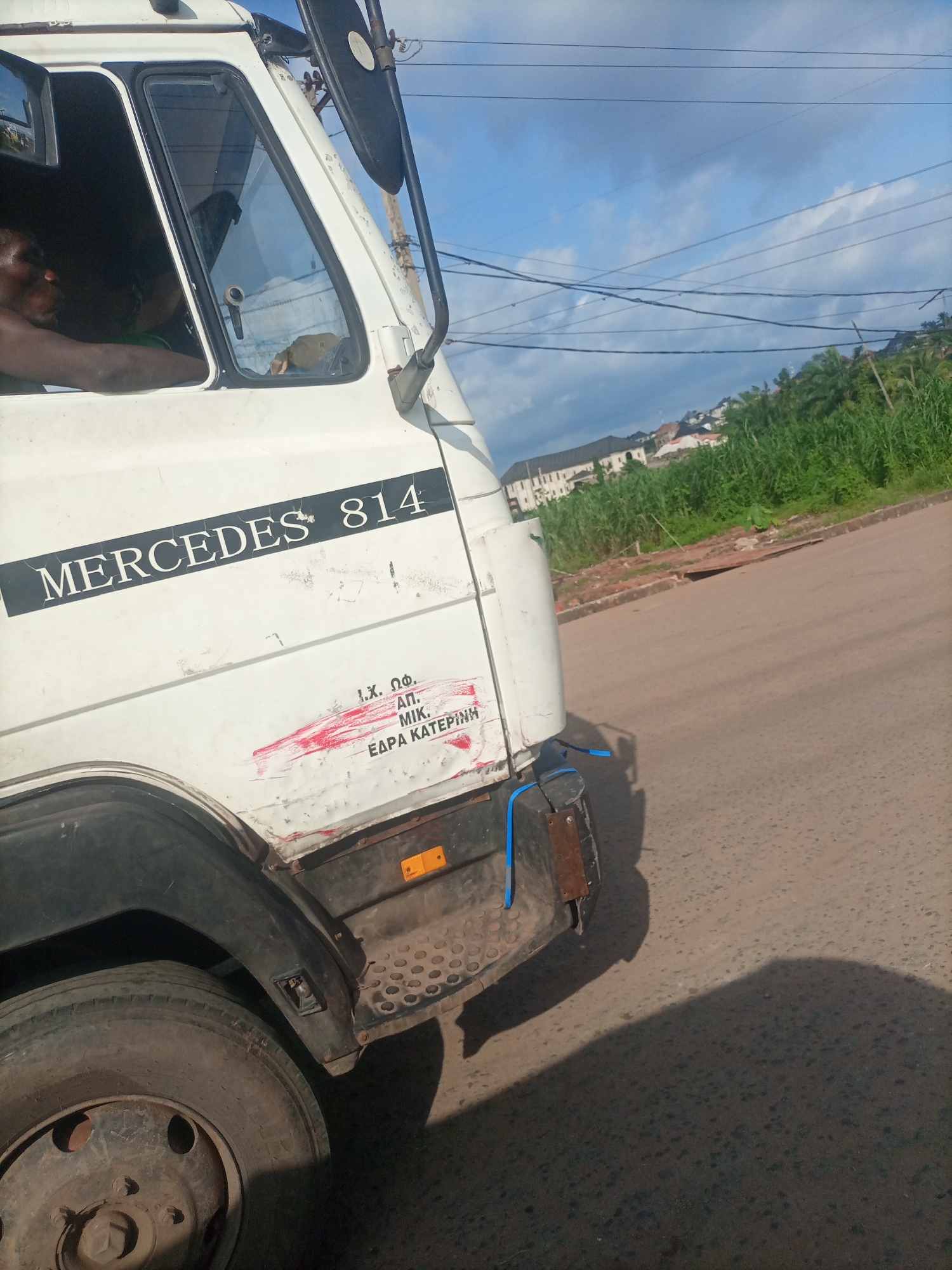 This is a medium shot of the side of a white Mercedes 814 truck, seen from a low angle on a paved road. The truck is white with black lettering that reads "MERCEDES 814". There are some red markings and what appears to be text in Greek letters on the side of the truck, partially obscured by a red smear. A man's head and arm are visible through the driver's side window, appearing to be resting. The truck is parked or moving slowly. In the background, there is lush green vegetation on a hillside with some buildings visible at the top. The sky is blue with scattered clouds, suggesting it is daytime, likely late afternoon or early morning due to the quality of light and shadows. The overall scene appears to be in a rural or semi-urban setting in Nkpor, Nigeria, indicated by the vegetation and the style of buildings in the distance. The weather seems fair.