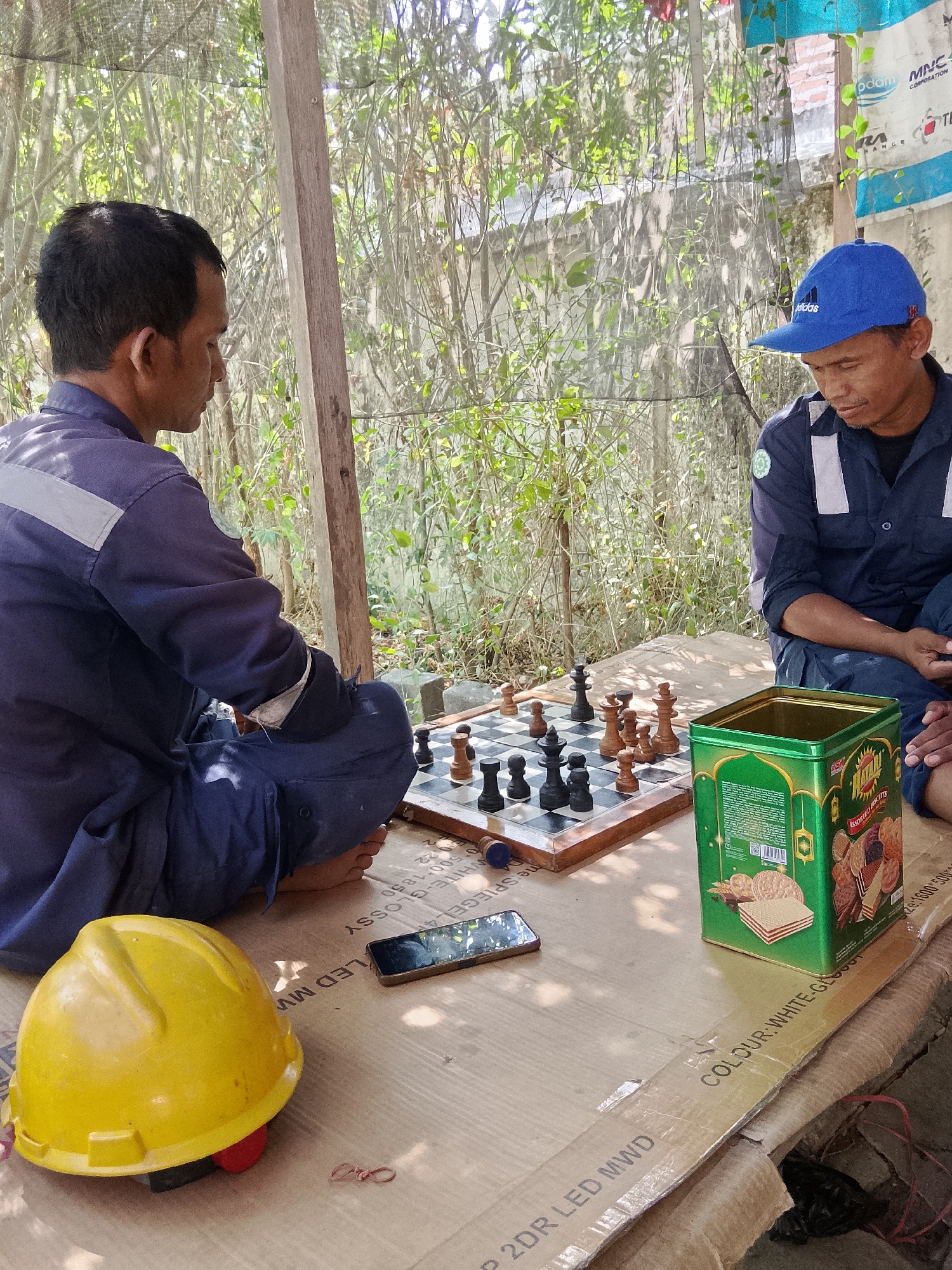 In the city of Tlogobendung, Indonesia, two men are engaged in a game of chess outdoors, seated on the ground. The man on the left, wearing a dark blue uniform with a grey reflective stripe on the shoulder, is facing away from the camera. The man on the right, also in a dark blue uniform, wears a blue Adidas baseball cap and is looking down at the chessboard. The chessboard is set up on a large piece of cardboard. Beside the chessboard is a green tin of "Matari Assorted Biscuits." A yellow hard hat lies on the cardboard to the left of the chessboard. The background consists of dense vegetation and trees, suggesting a natural or semi-rural setting. The scene is lit by natural daylight.