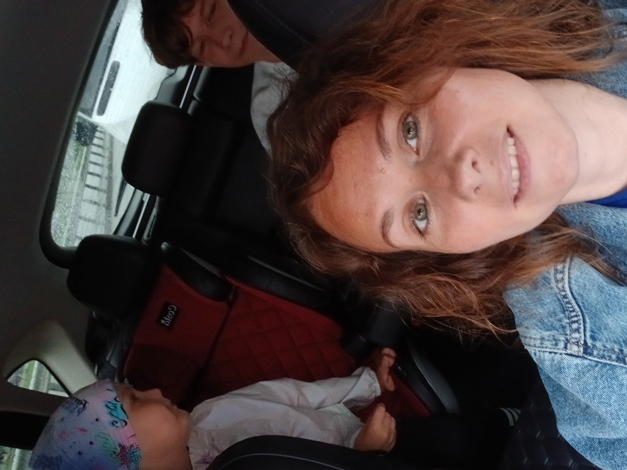 The image is a selfie taken inside a car, presumably on a rainy day as evidenced by the blurred windshield and the water droplets on the glass. The foreground features a woman with brown hair and green eyes, wearing a denim jacket. To her left, a young child with a colorful headscarf is visible, looking to the side. In the background, another person, likely a teenager, is partially visible behind a car seat. The car's interior includes red and black upholstery and a child car seat. The environment outside the car appears to be a road with traffic. There are no specific Yekaterinburg landmarks visible in the image.