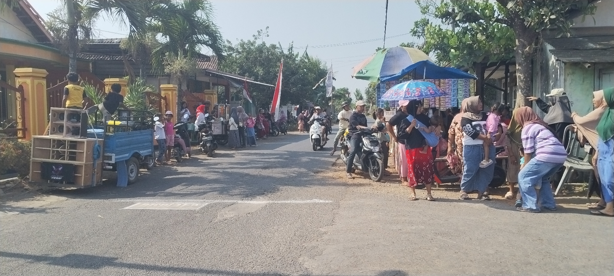 In Yosowilangun Kidul, Indonesia, on a sunny day, a street scene unfolds with a lively gathering of people. The image captures a small truck adorned with a large sound system, positioned on the left side of the road. Two young men are visible on the truck, one wearing a yellow shirt and the other a black shirt, appearing to be operating or setting up the equipment. Several children and adults are standing on the roadside, some near motorcycles.
Further down the street, a makeshift stall with colorful umbrellas is set up, displaying various items for sale. A group of people are gathered around this stall, engaged in what appears to be a social event or local market activity. Indonesian flags are visible in the background, suggesting a celebratory or national occasion. The environment is characterized by residential buildings and trees lining the road, indicative of a village or small town setting. The road surface is asphalt, with a white line marking the center. Shadows cast by the sun and foliage suggest it is daytime.