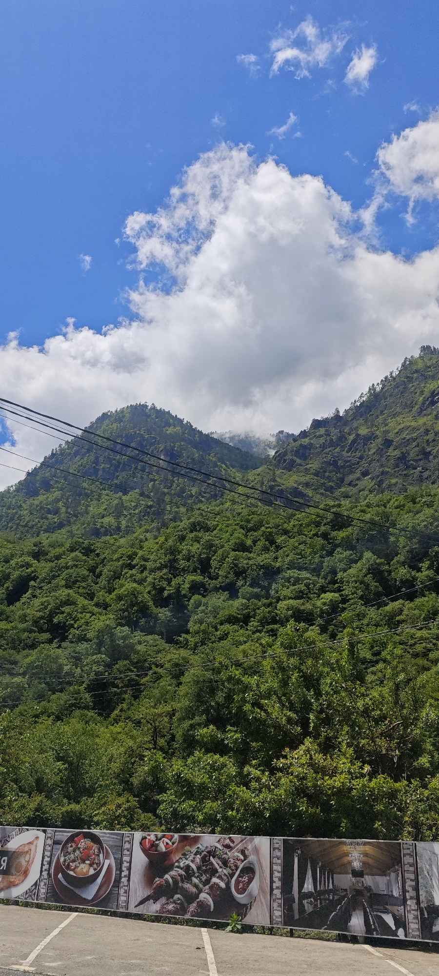 The image displays a vibrant mountainous landscape under a bright blue sky dotted with white cumulus clouds. Dense, lush green forests cover the steep slopes of the mountains, which dominate the upper two-thirds of the frame. Towards the distant peaks, a soft mist or lower clouds gently cling to the mountainsides.
Several dark power lines or cables stretch across the mid-ground, providing a visual element of human infrastructure against the natural backdrop. No people are visible in the scene.
The time of day is clearly daytime, with bright, direct lighting suggesting mid-day. The weather is fair, with blue skies and picturesque clouds, though the mist on the distant mountains hints at potential humidity or lingering cloud cover.
At the bottom of the image, a horizontal banner or billboard is prominently featured, advertising a restaurant or food establishment. From left to right, the banner sections show:
1. A dish resembling a type of pastry or bread (possibly Khachapuri), with partially visible Cyrillic text next to it.
2. A bowl filled with a savory-looking stew or mixed dish.
3. Multiple skewers of grilled meat (like shashlik or kebabs) presented on a wooden board, accompanied by sauces and a small salad.
4. An interior view of a dining hall or restaurant, featuring long tables and rustic décor.
The scene combines natural beauty with signs of human activity, characteristic of a scenic area with amenities for travelers or locals, fitting the location context of Bzypta, Abkhazia.