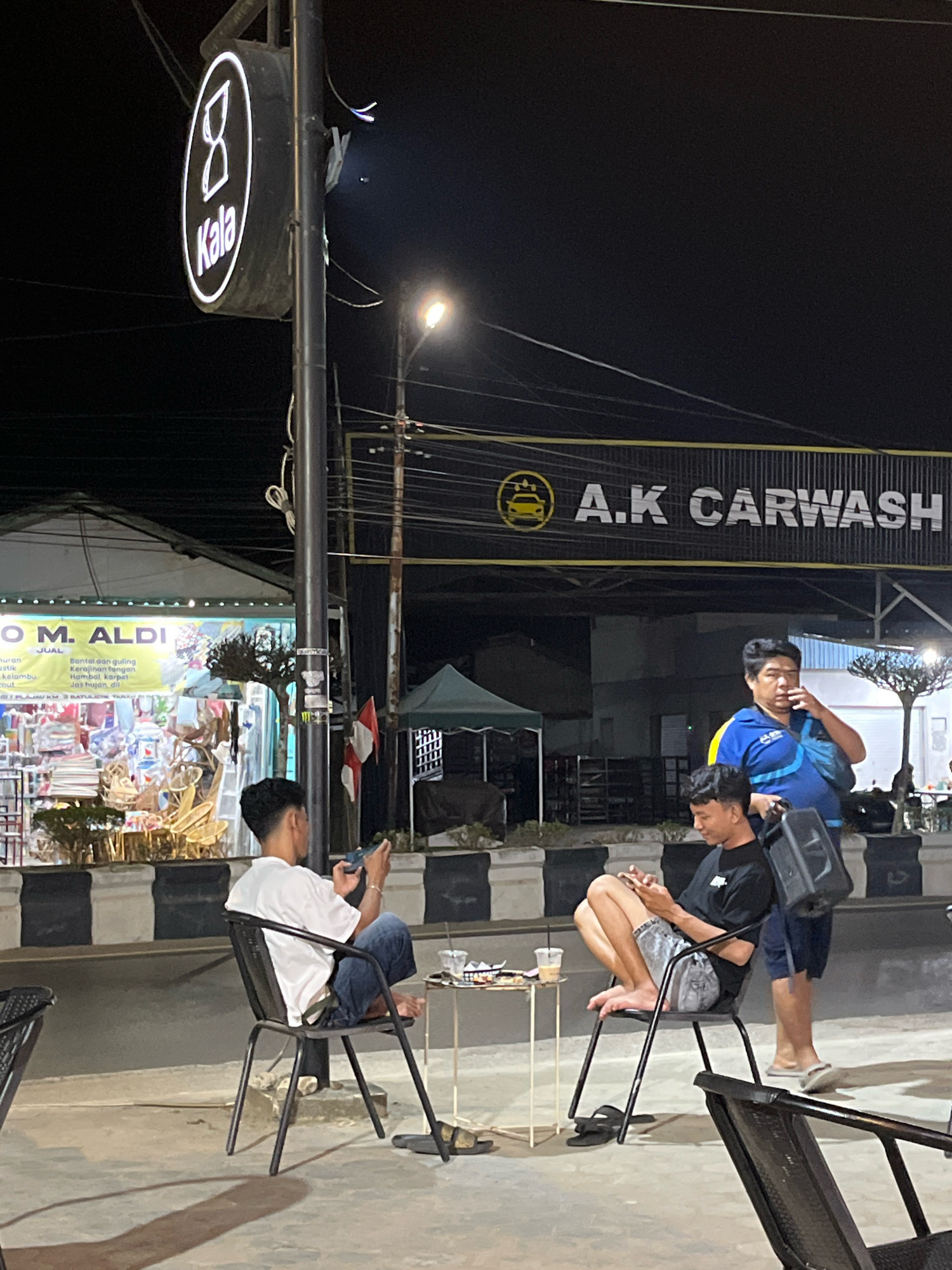 In Plajau Mulia, Indonesia, at night, a scene unfolds outside a "KALA" branded establishment, possibly a cafe or restaurant, adjacent to an "A.K CARWASH". Three individuals are present. One man, wearing a blue shirt with blue and yellow accents and a black strap across his chest, stands near a black speaker, holding his hand to his mouth as if speaking or thinking. To his left, a young man in a white t-shirt and blue jeans sits on a black metal chair, looking down at his phone. Another young man, seated on a similar chair, wears a black t-shirt and grey shorts and is also engrossed in his phone. A small white table with drinks and what appears to be food is situated between them. In the background, a storefront with a sign reading "O M. ALDI" displays various goods. The environment is lit by streetlights and illuminated signs, creating a nighttime ambiance.