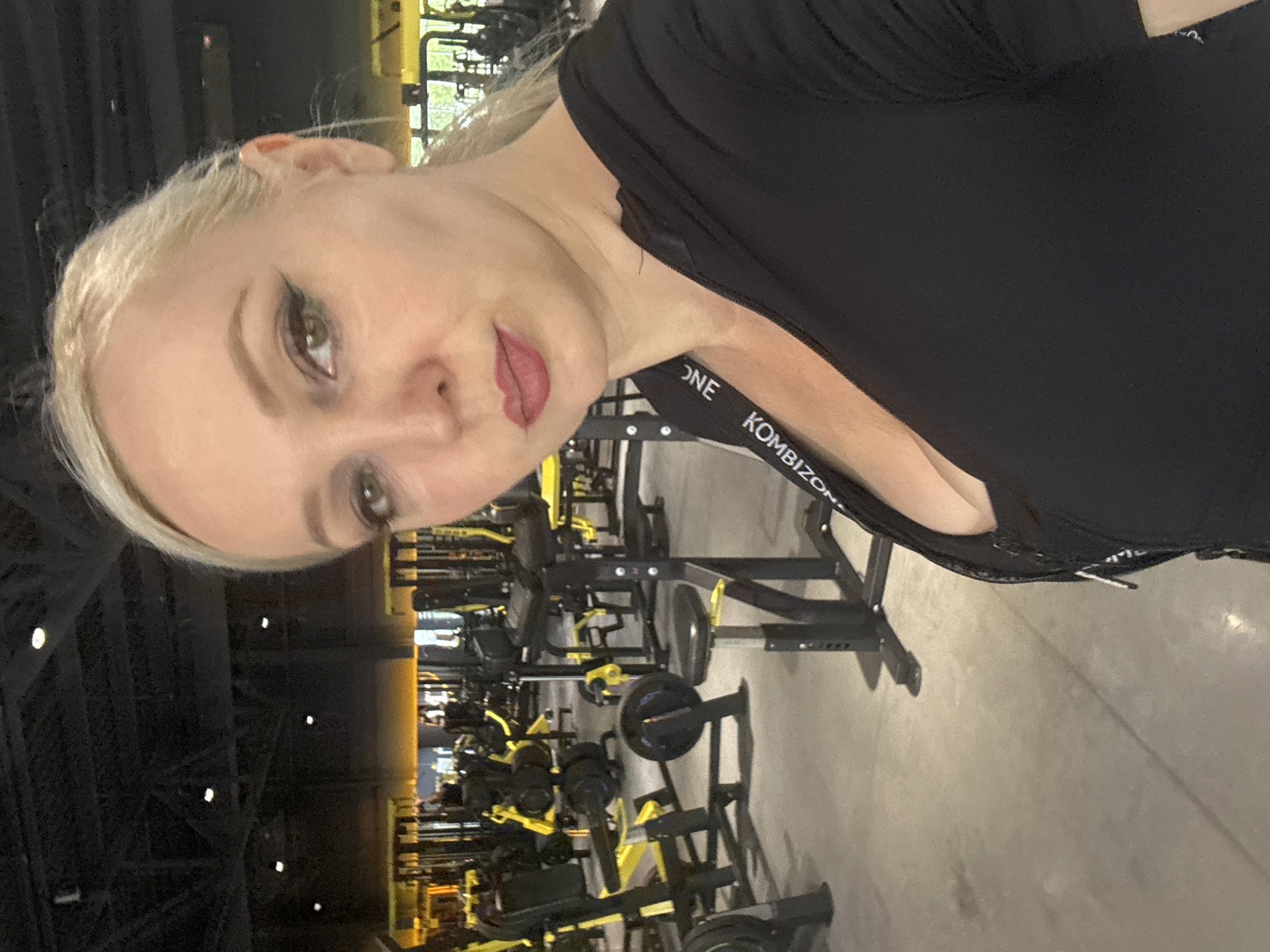 A blonde woman with winged eyeliner and red lipstick takes a selfie in a gym. She is wearing a black zip-up top with a black and white ribbon that reads "KOMBISON" visible on the strap. The gym is filled with yellow and black weight machines and equipment. The floor is a light gray concrete. The ceiling is black and appears to have exposed pipes and lighting fixtures. The woman is positioned in the foreground, facing the camera, with the gym equipment in the background. The scene is well-lit. The image was taken indoors.
