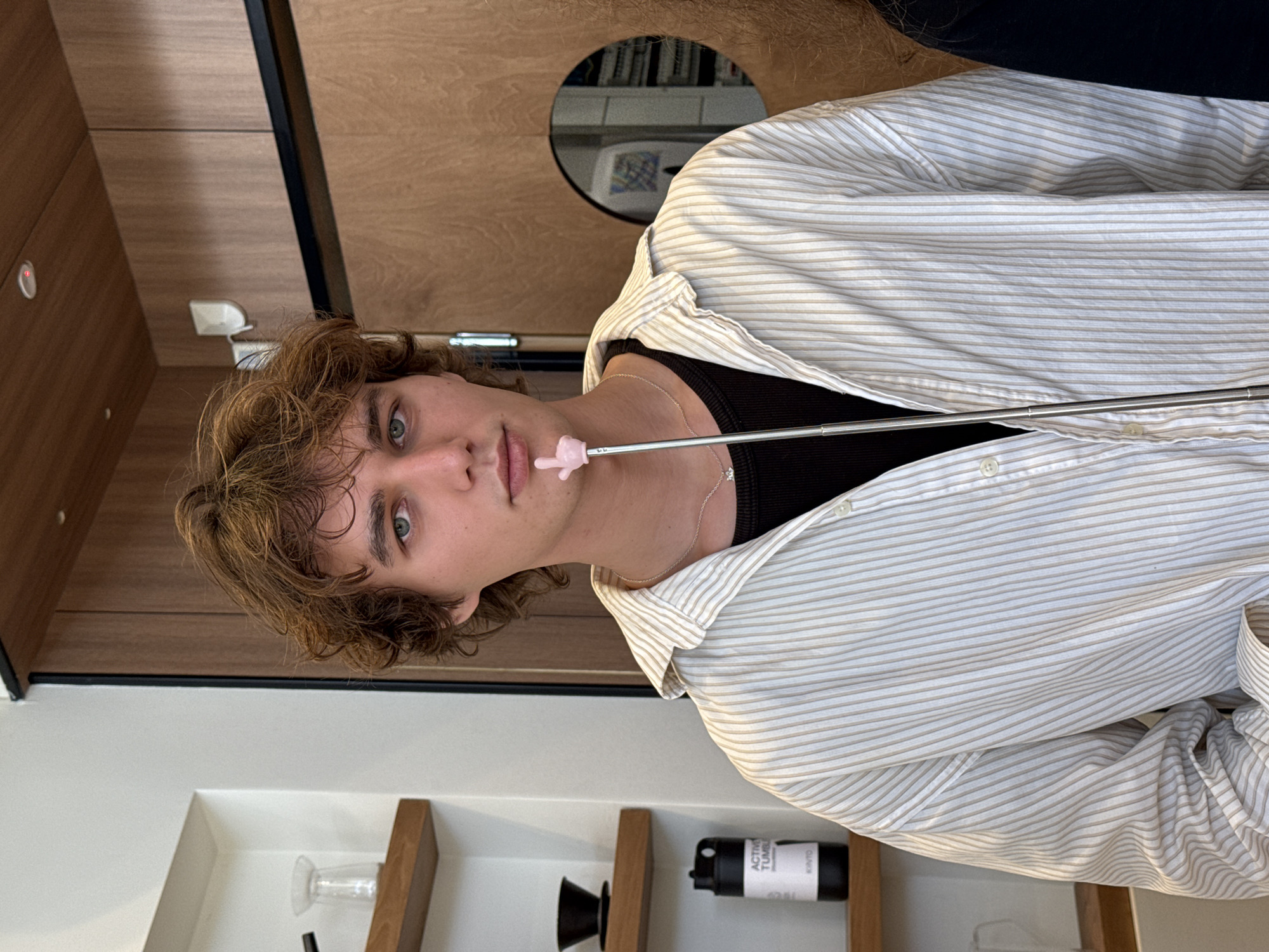 A person with light brown, wavy hair, wearing a black tank top and a light-colored striped button-down shirt, is positioned in front of a wooden wall. The person has a pink object on the end of a thin metal rod near their mouth. The environment includes shelves with various items, such as a glass, a coffee filter, and a black canister labeled "Active Tumbler KINTO". A circular opening in the wooden wall is visible in the background. The scene is likely indoors. The image's orientation is rotated 90 degrees clockwise. There is no verifiable evidence the image was taken in Tel Aviv, Israel.
