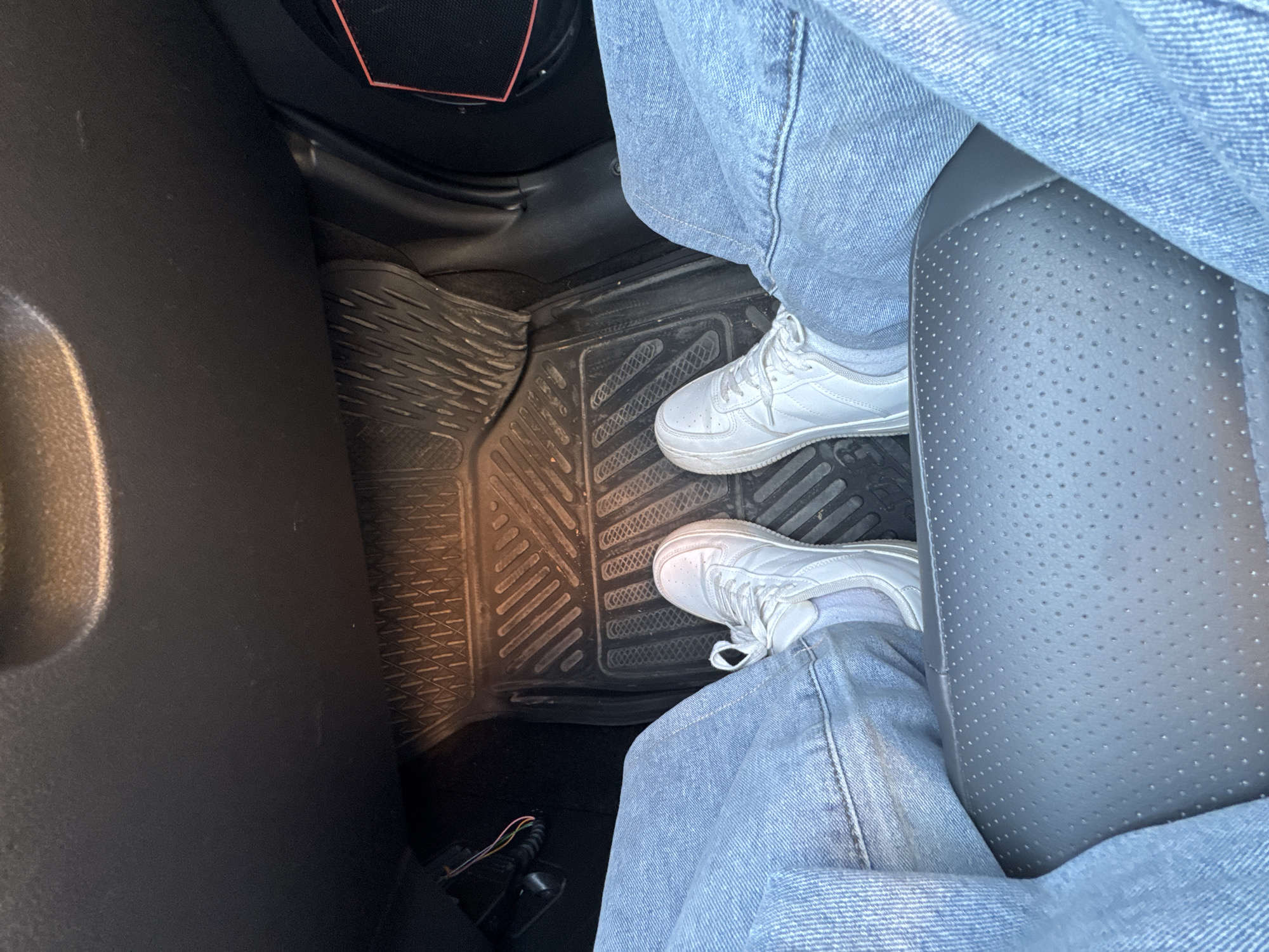 A person wearing light-wash denim jeans and white sneakers is seated in the passenger seat of a vehicle. The vehicle has black rubber floor mats and perforated gray leather seats. The image was likely taken indoors, inside of the vehicle. The location within the city of Perm, Russia is indeterminable.