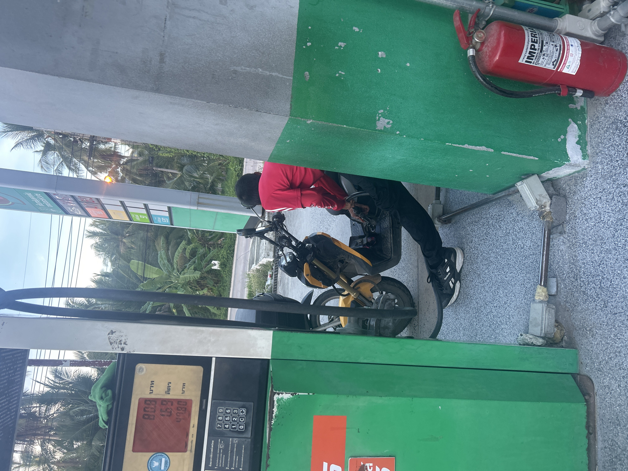 Here is a detailed description of the image:
**Location:**
* The image appears to be taken at a gas station in ตำบลเกาะพะงัน, Thailand. Palm trees are visible in the background, and the pump displays Thai text and currency units.
**People:**
* One person is visible. They are wearing a red long-sleeved shirt and black pants. They are positioned next to a yellow motorcycle at the gas pump, appearing to be refueling it.
**Objects:**
* **Gas Pump:** A green and white gas pump dominates the foreground. The display shows "80.8" (possibly the price), "1.69" (likely the liters pumped), and "47.80" (the total cost). A green cloth is draped over the top of the pump.
* **Motorcycle:** A yellow and black motorcycle is parked next to the gas pump.
* **Fire Extinguisher:** A red "IMPERIA" fire extinguisher is mounted on the wall adjacent to the gas pump.
**Actions and Interactions:**
* The person is interacting with the motorcycle, presumably to refuel it.
**Environment:**
* The setting appears to be a typical outdoor gas station environment. The ground is paved and speckled. There is lush vegetation, including palm trees, in the background, indicating a tropical climate. The lighting suggests it is daytime, possibly late afternoon.
