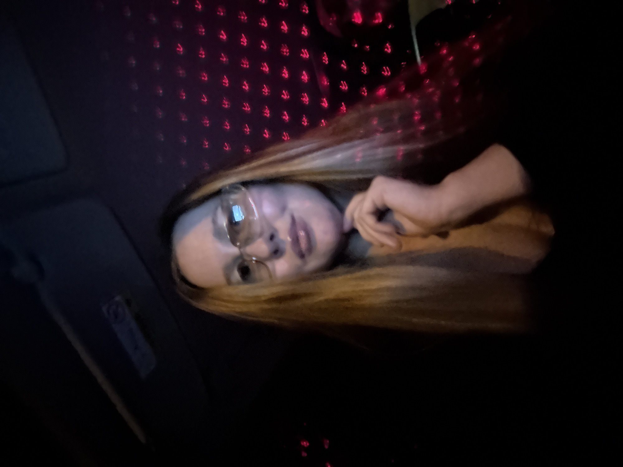 A young woman with long blonde hair and glasses is in the driver's seat of a vehicle at night. She is looking at the camera with a slight smile, her hand resting on her chin. The interior of the car is dimly lit, with red lights reflecting off the windshield and the woman's face. The background shows a pattern of small red lights, possibly from a decorative lighting system or a projection. The image appears to have been taken in the city of Chaikovsky, Russia, as indicated by the metadata.